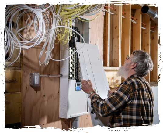 A man with gray hair and glasses in a plaid shirt working on an electrical panel in a workshop with exposed wood framing and hanging electrical wires.