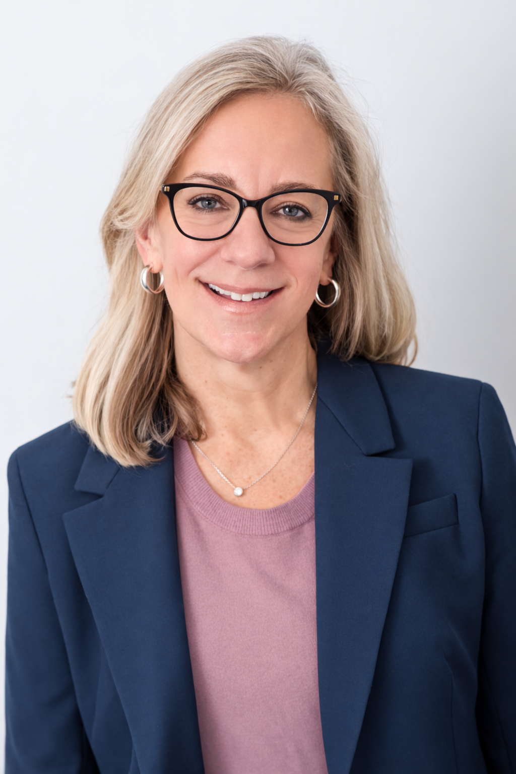 A woman with blonde hair, wearing glasses, hoop earrings, a navy blazer, and a pink top, smiling at the camera.