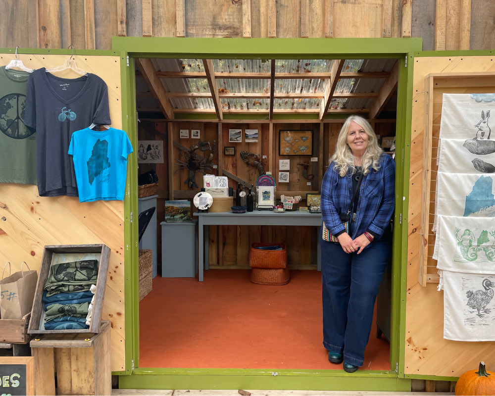 A woman standing in the entrance of a rustic shop with handmade art and apparel, including t-shirts with nature-themed prints, in a wooden setting with a sloped, translucent roof.