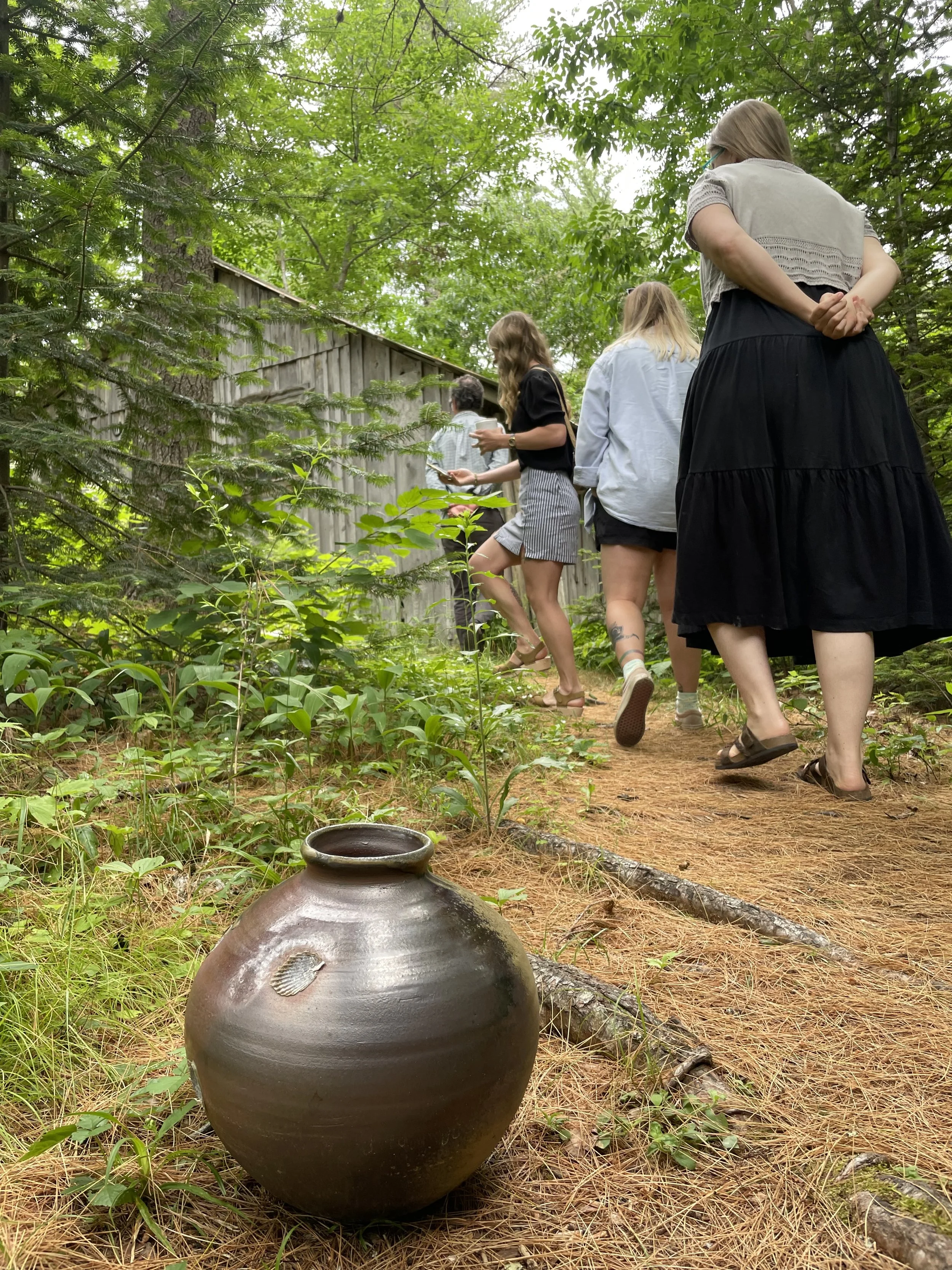 A group of women walking along a wooded forest trail, seen from behind, with lush green foliage and a wooden fence on the side. In the foreground, there is a brown ceramic pot on the ground.