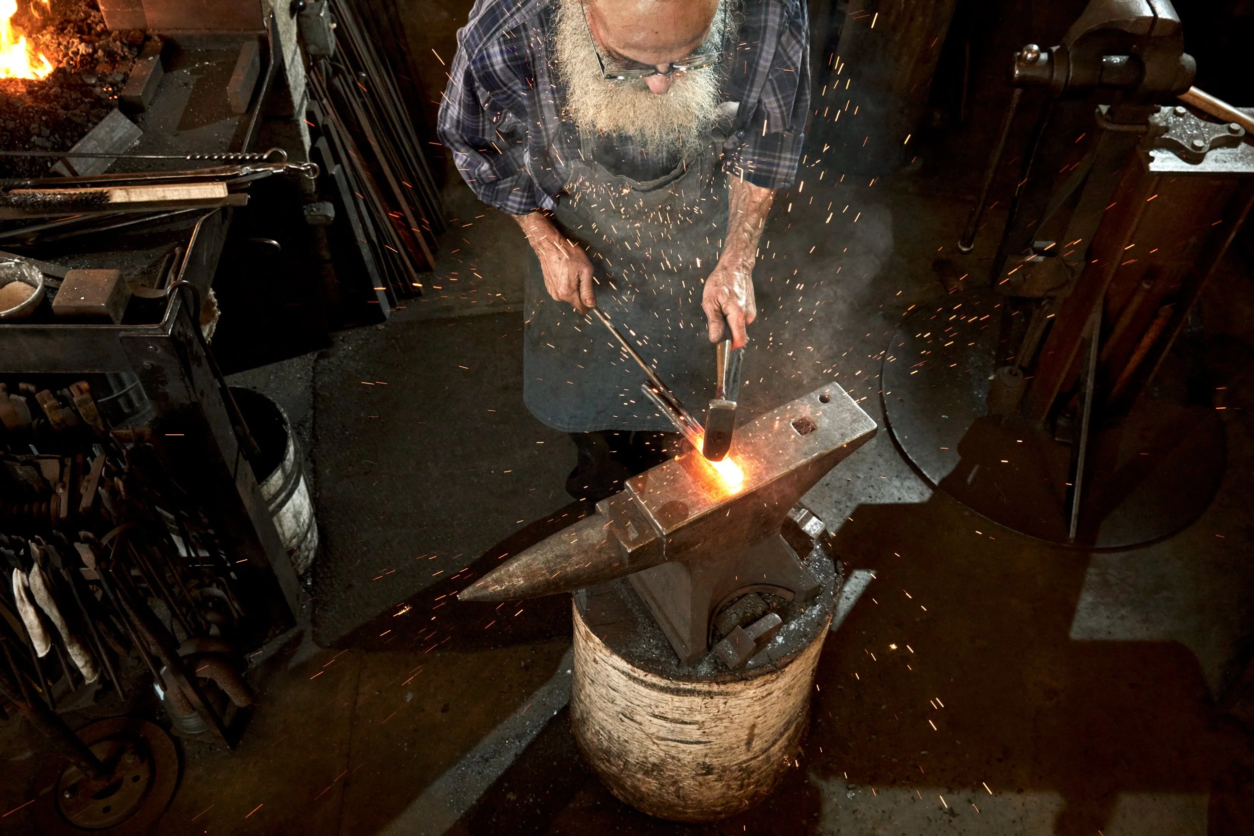An older man with a beard and glasses working at an anvil, forging metal with sparks flying.