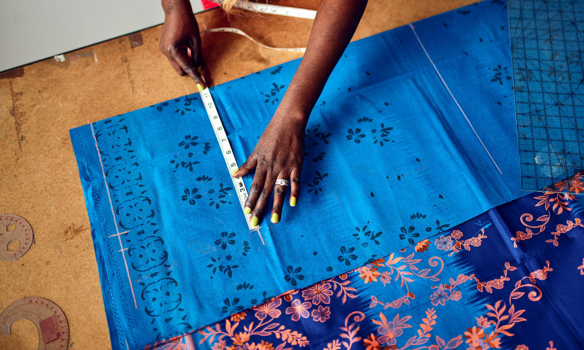 A person with lime green nail polish measuring vivid blue fabric with floral patterns using a measuring tape at a worktable.