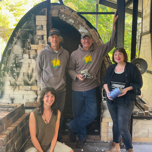 Four people posing outdoors in front of a brick kiln, two men wearing gray sweatshirts with a yellow logo, two women, one sitting and one holding a cup, smiling at the camera.