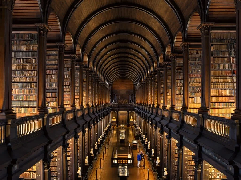A grand, two-story library interior with high vaulted ceilings, tall wooden bookshelves filled with books, and a walkway with a railing down the center. Several visitors are browsing and reading.