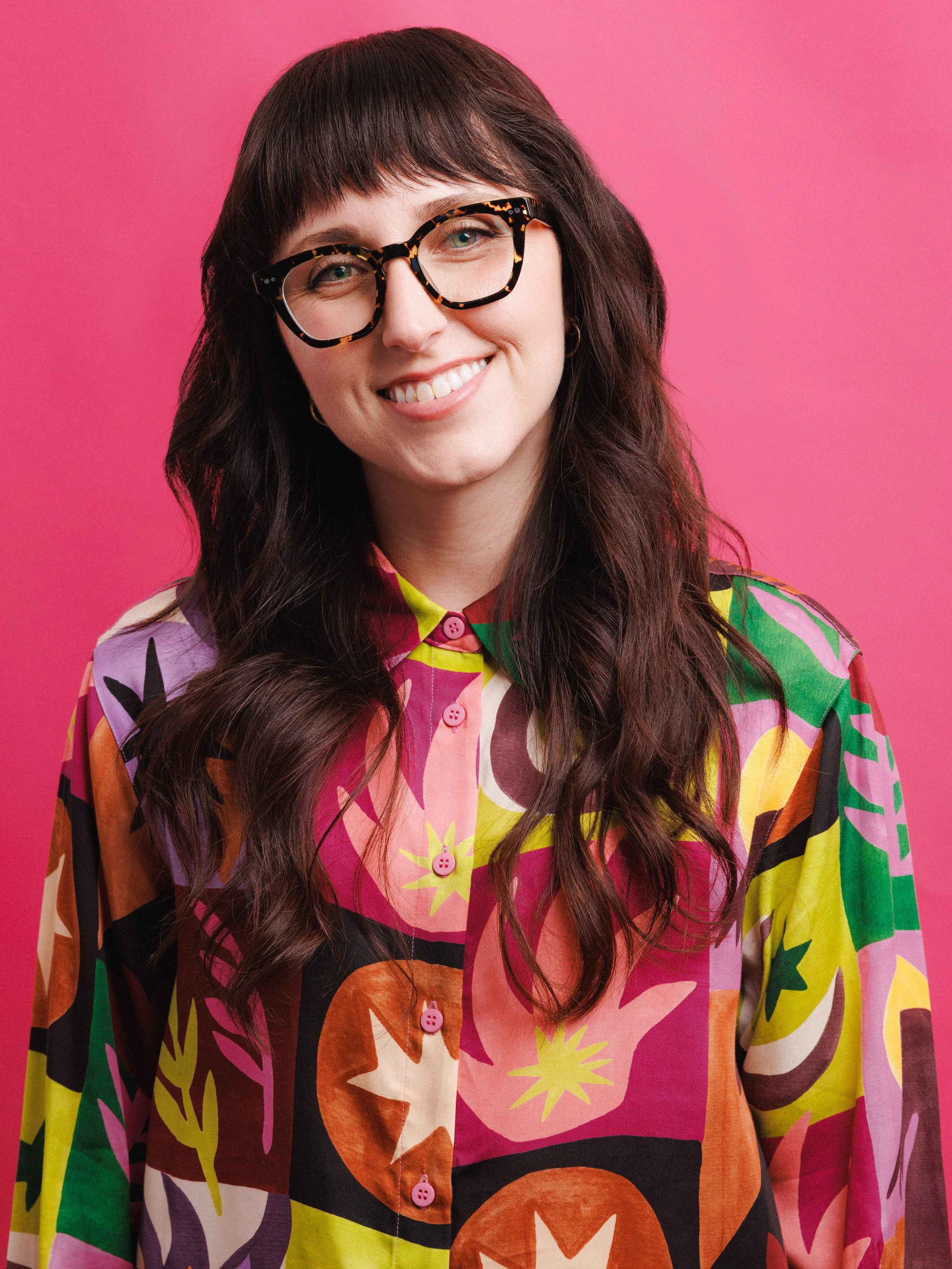 A photo of Natalie Rempalski, smiling against a pink background, wearing a multicolored, patterned shirt, framed by both her bold glasses and long, dark brunette, wavy hair and bangs.