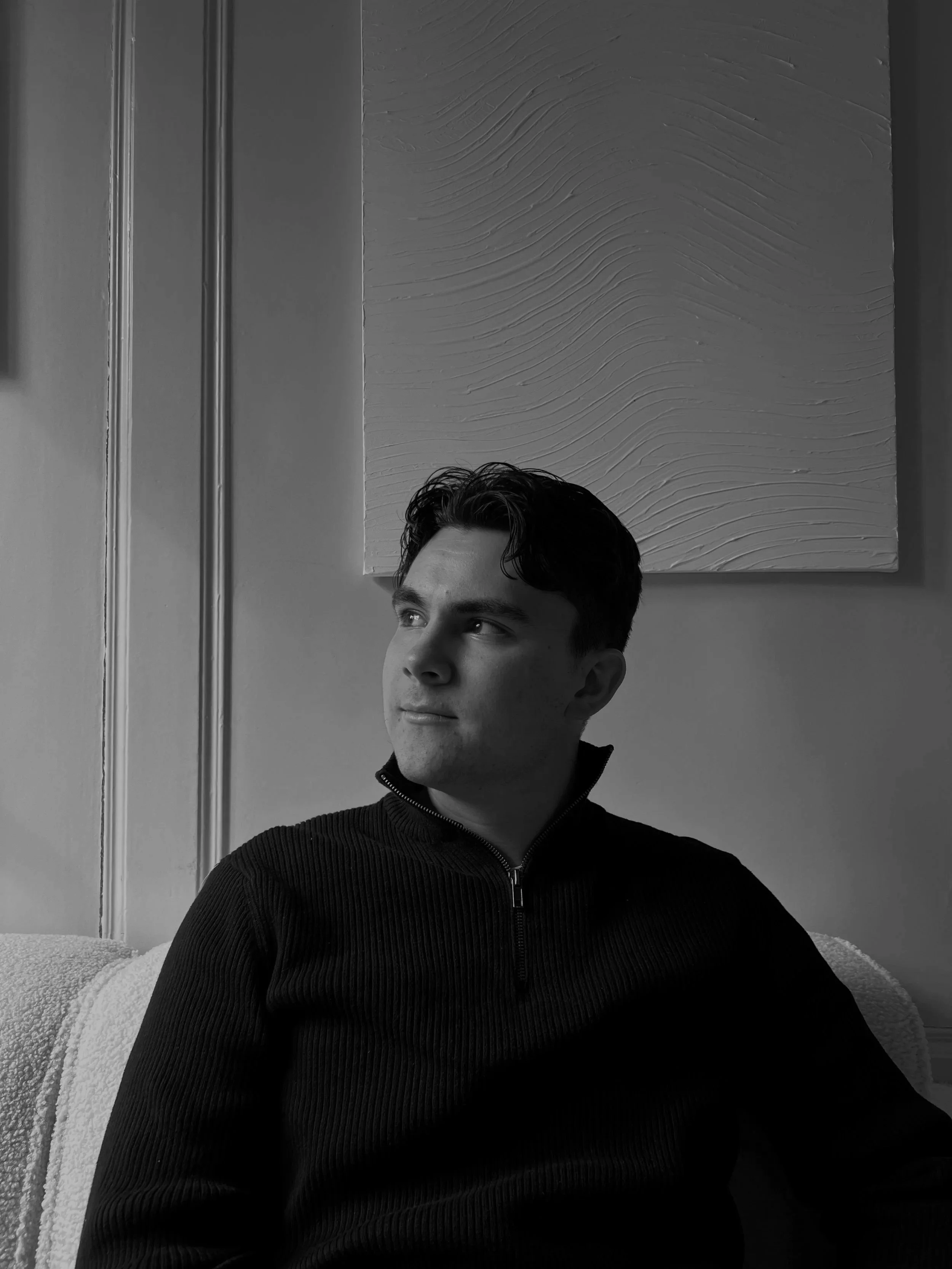 Ein junger Mann sitzt auf einem Sofa, blickt nach links, trägt einen schwarzen Pullover mit Reißverschluss, im Hintergrund eine abstrakte weiße Wanddekoration.