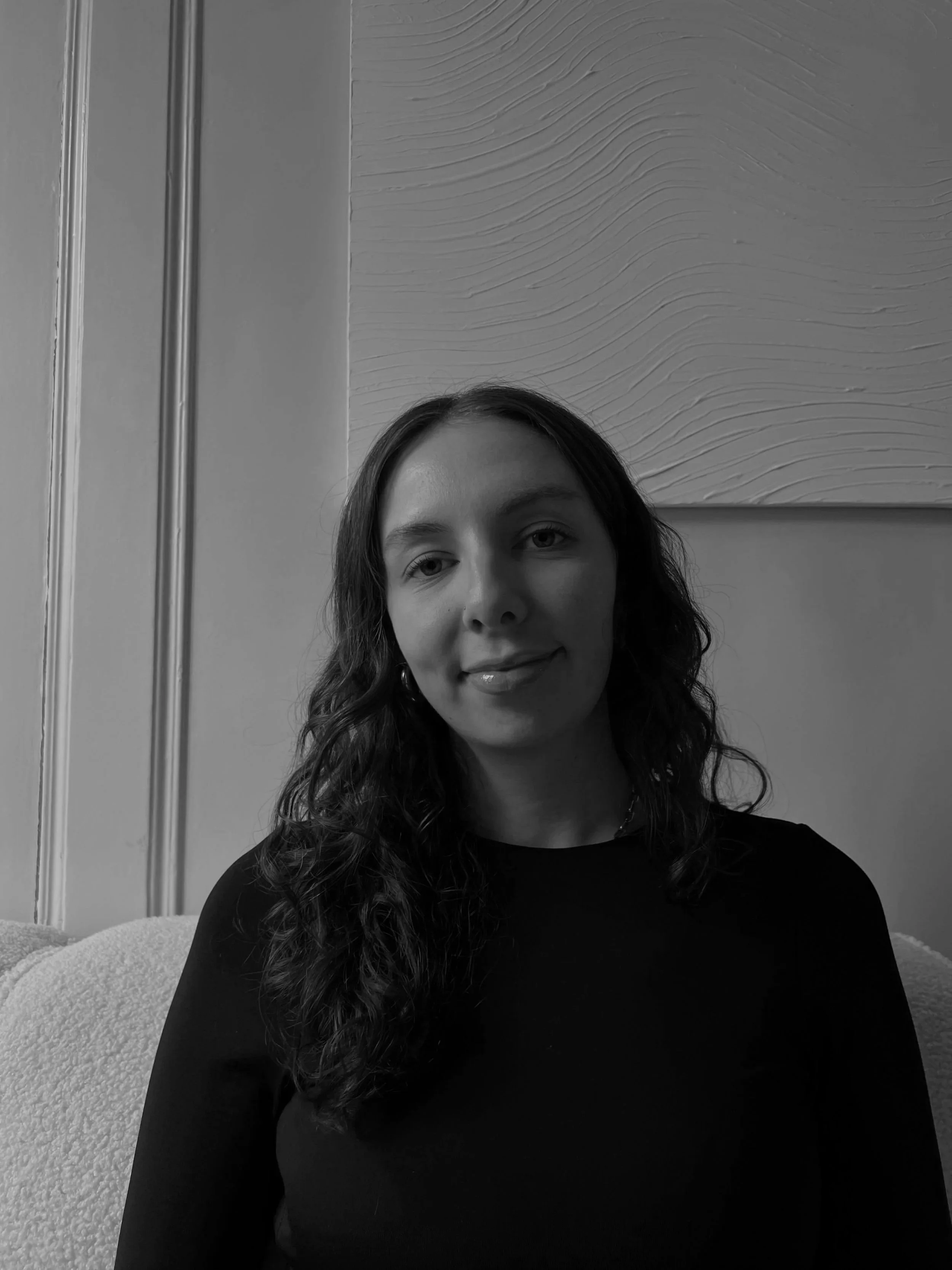 Schwarz-weiß-Foto eines lachenden jungen Mädchens mit langen, gewellten Haaren, das auf einem Sofa sitzt, im Hintergrund Wand mit Textur.