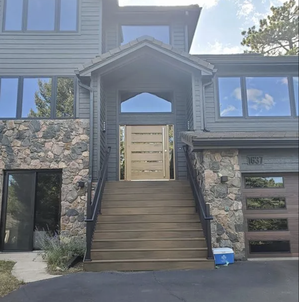 Front view of a modern Evergreen, Colorado house with stone and Diamond Kote siding, elevated by wooden stairs with black railings leading to a closed wooden gate. House has large Andersen 100 Series windows reflecting the sky and trees.