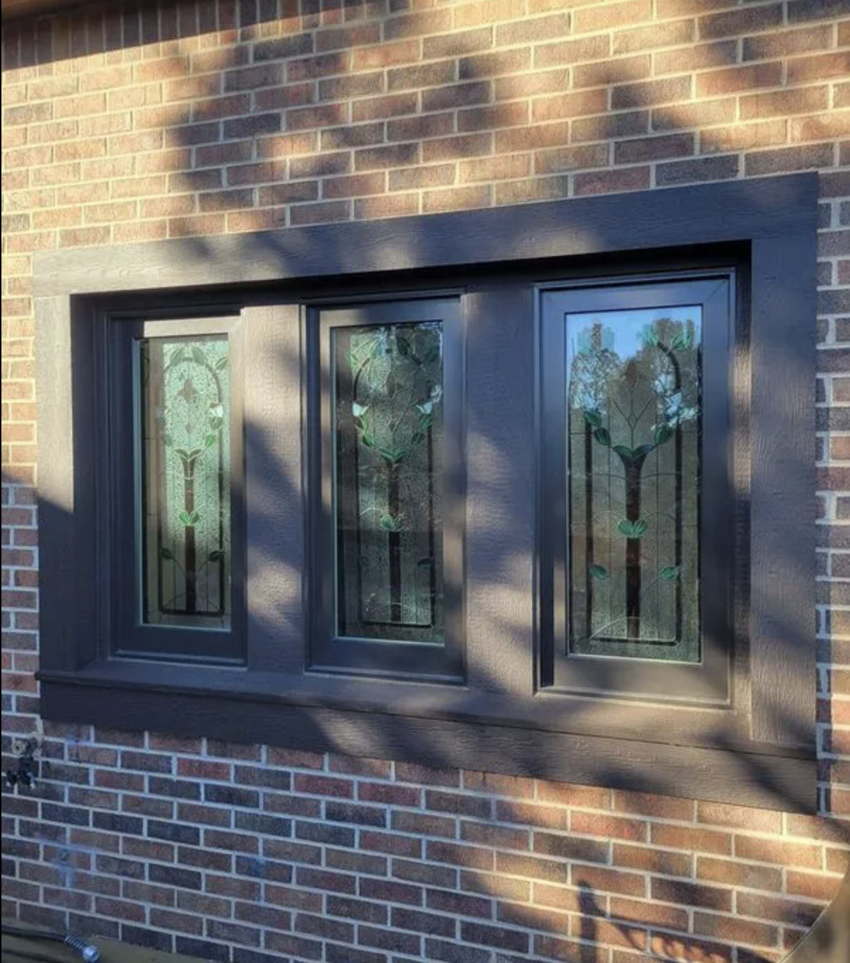 A decorative Andersen A-Series window with three stained glass panels featuring green floral designs, set in a black frame on a brick wall in Colorado Springs, Colorado.