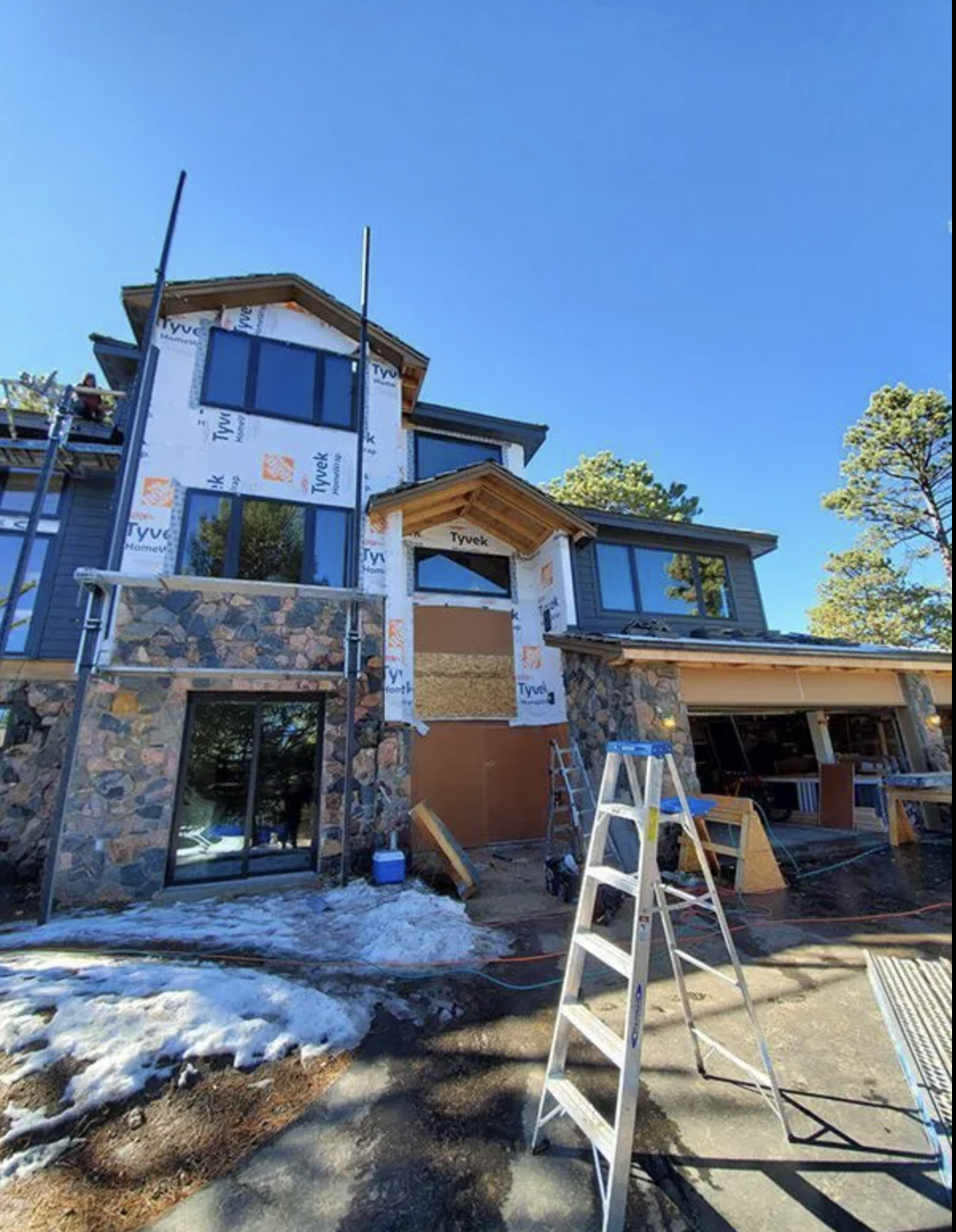Evergreen, Colorado house under construction with scaffolding and ladder, partially covered with Tyvek weather barrier, Diamond Kote siding, and construction materials on site, snow on ground, clear blue sky.