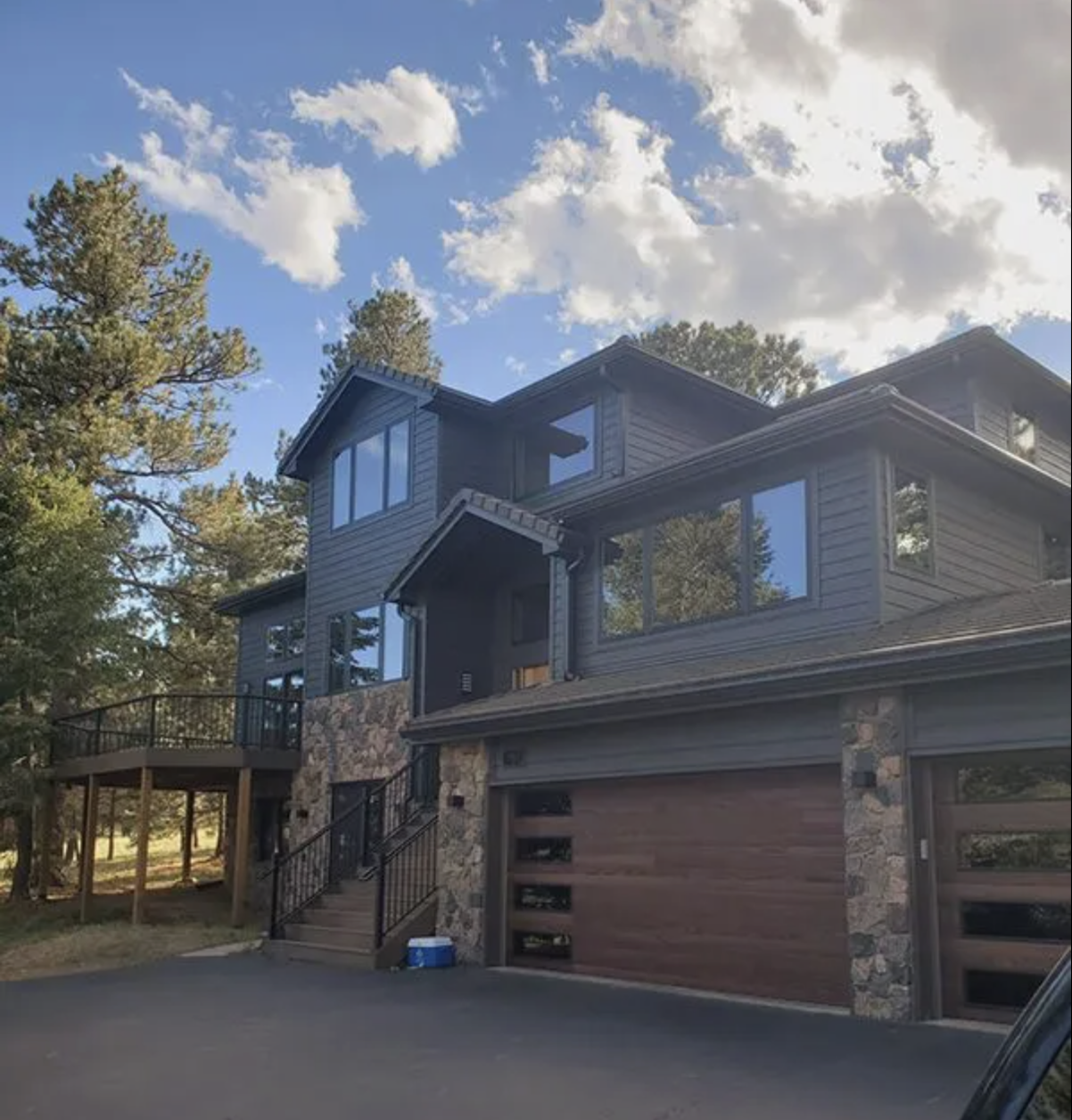 Modern multi-story Evergreen, Colorado house with large black Andersen 100 Series windows, Diamond Kote siding exterior, surrounded by tall trees, under a partly cloudy sky.
