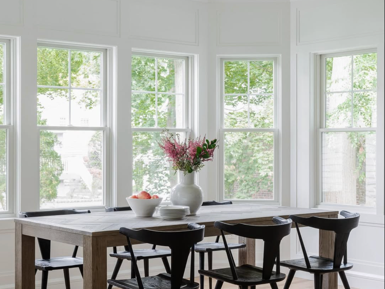 Bright dining room with a wooden table, black chairs, and a white vase with pink flowers, viewed through large white Andersen 400 Series windows showing green trees outside.