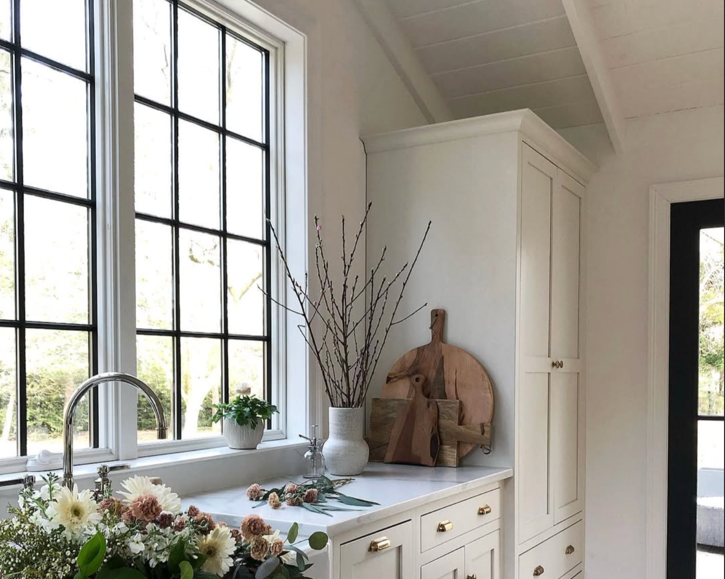 Kitchen interior with large white Andersen E-Series window, white cabinetry, decorative cutting boards, flowers, and a potted plant.