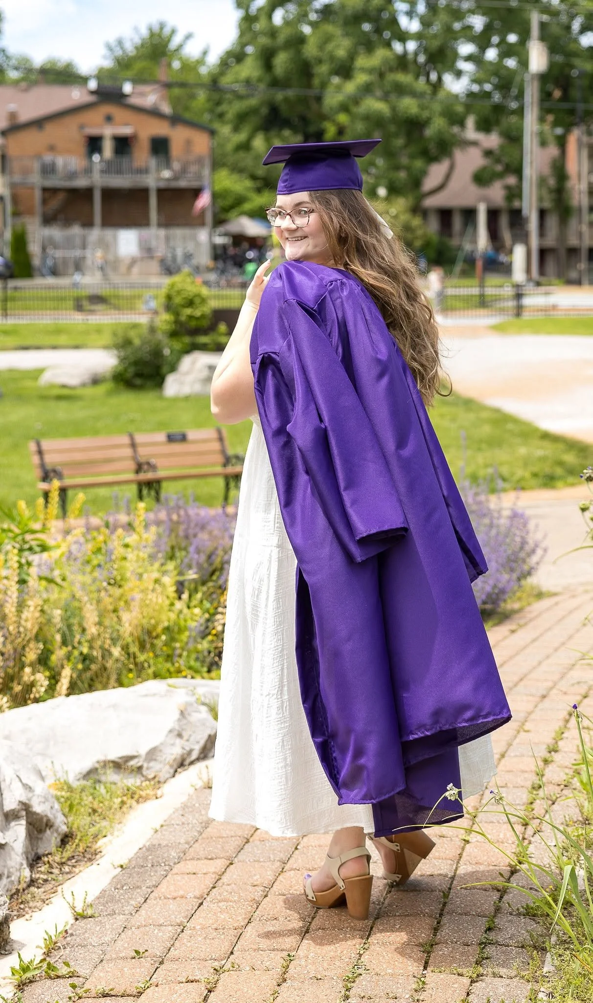 A young woman wearing glasses and a purple graduation gown with a matching cap, smiling while posing outdoors on a brick pathway with greenery and houses in the background.