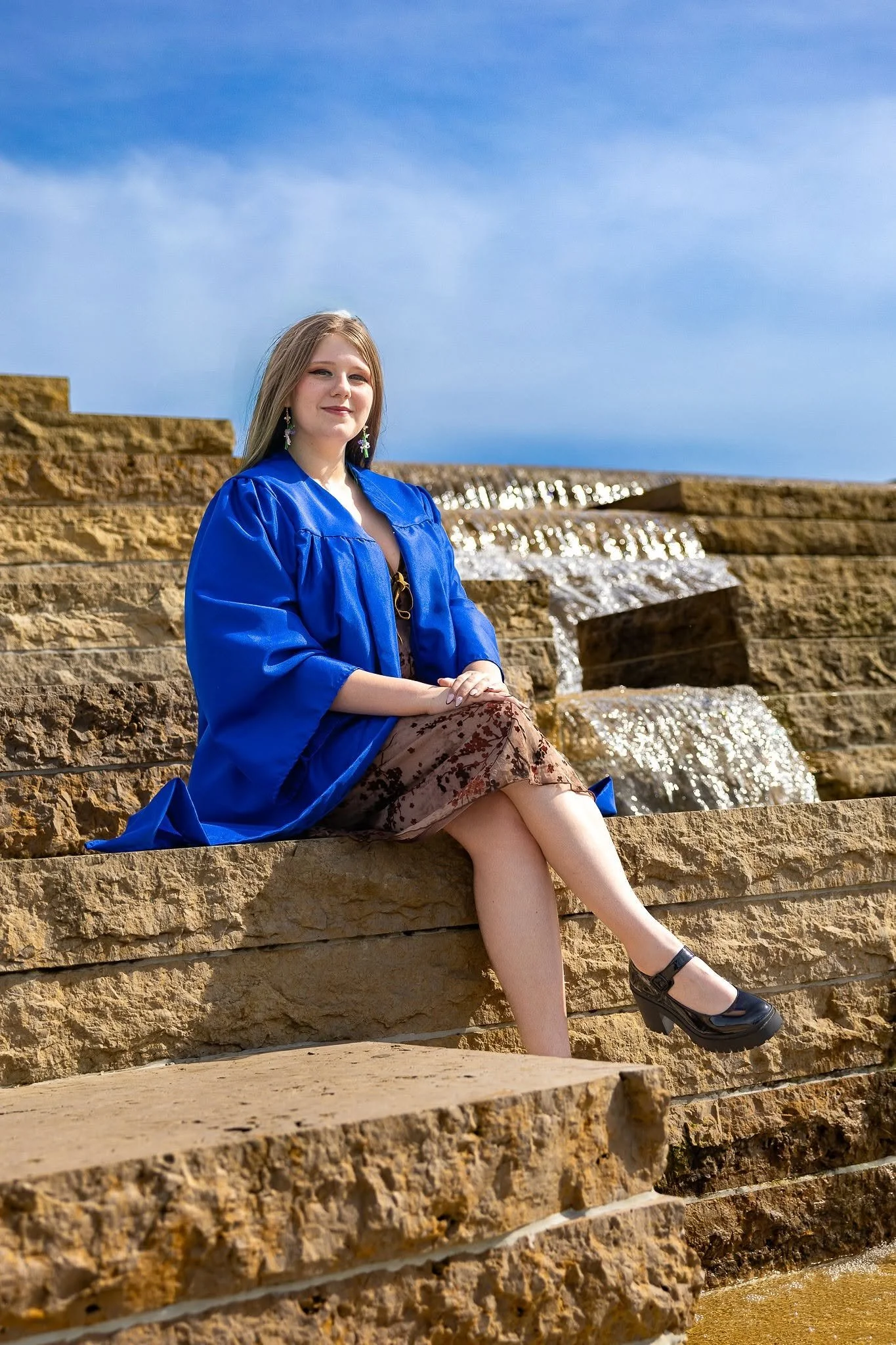 A young woman wearing a blue graduation gown sitting on stone steps with a waterfall and blue sky in the background.