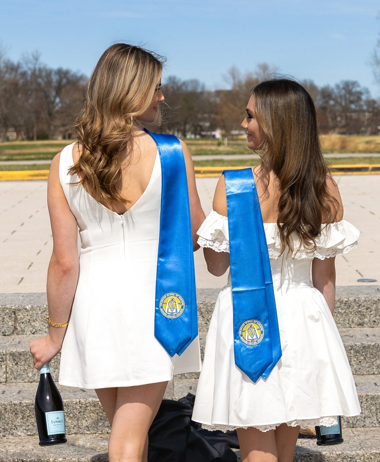 Two women in white dresses with blue sashes standing outdoors, holding bottles of La Marca wine, and smiling at each other.