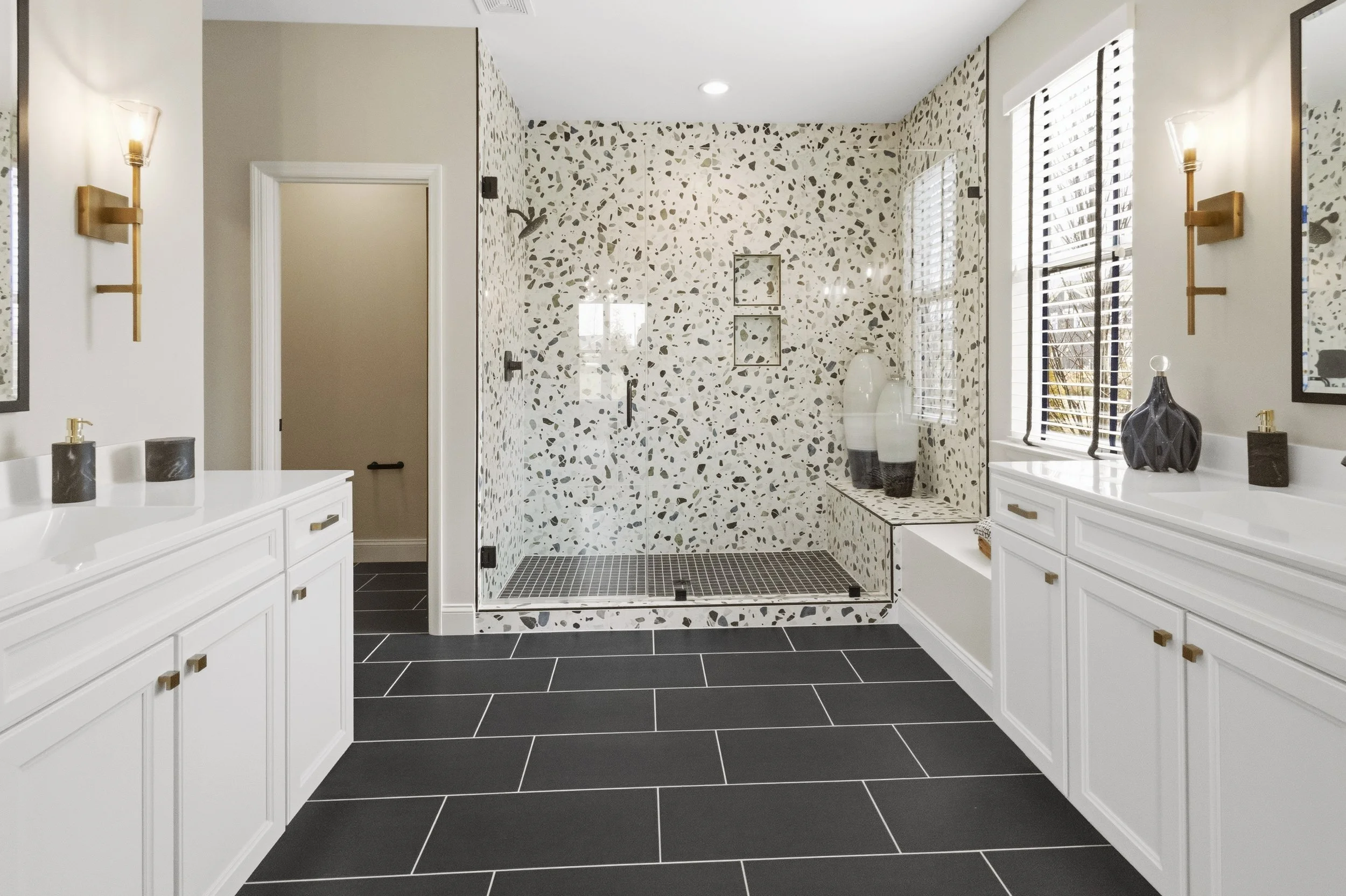 Modern bathroom with white vanities, gold hardware, black soap dispensers, a walk-in shower with terrazzo tile, black floor tiles, and a large window with white blinds.