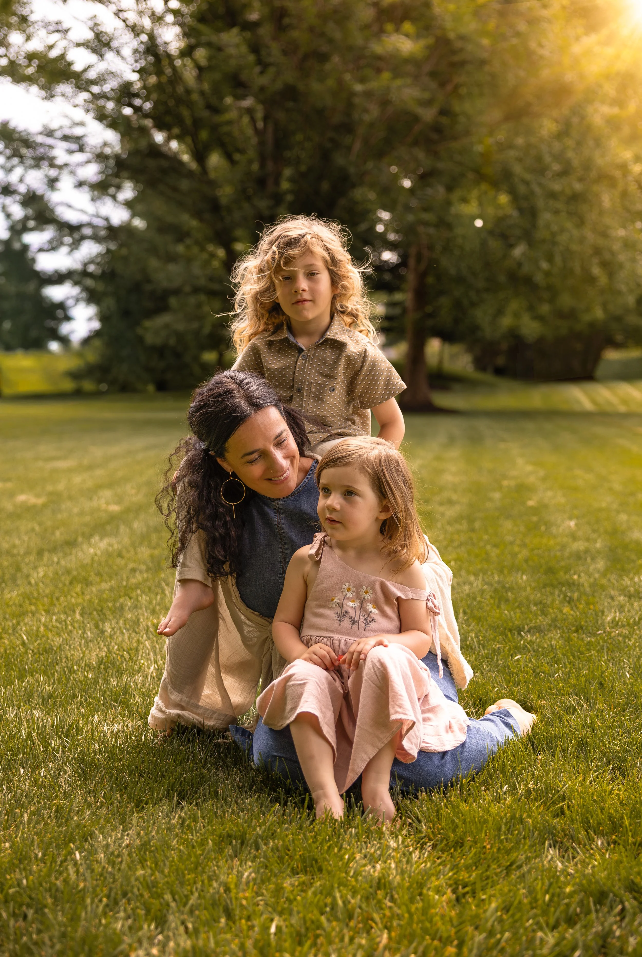 A woman and three young girls sitting on the grass in a park during sunset, with trees in the background.