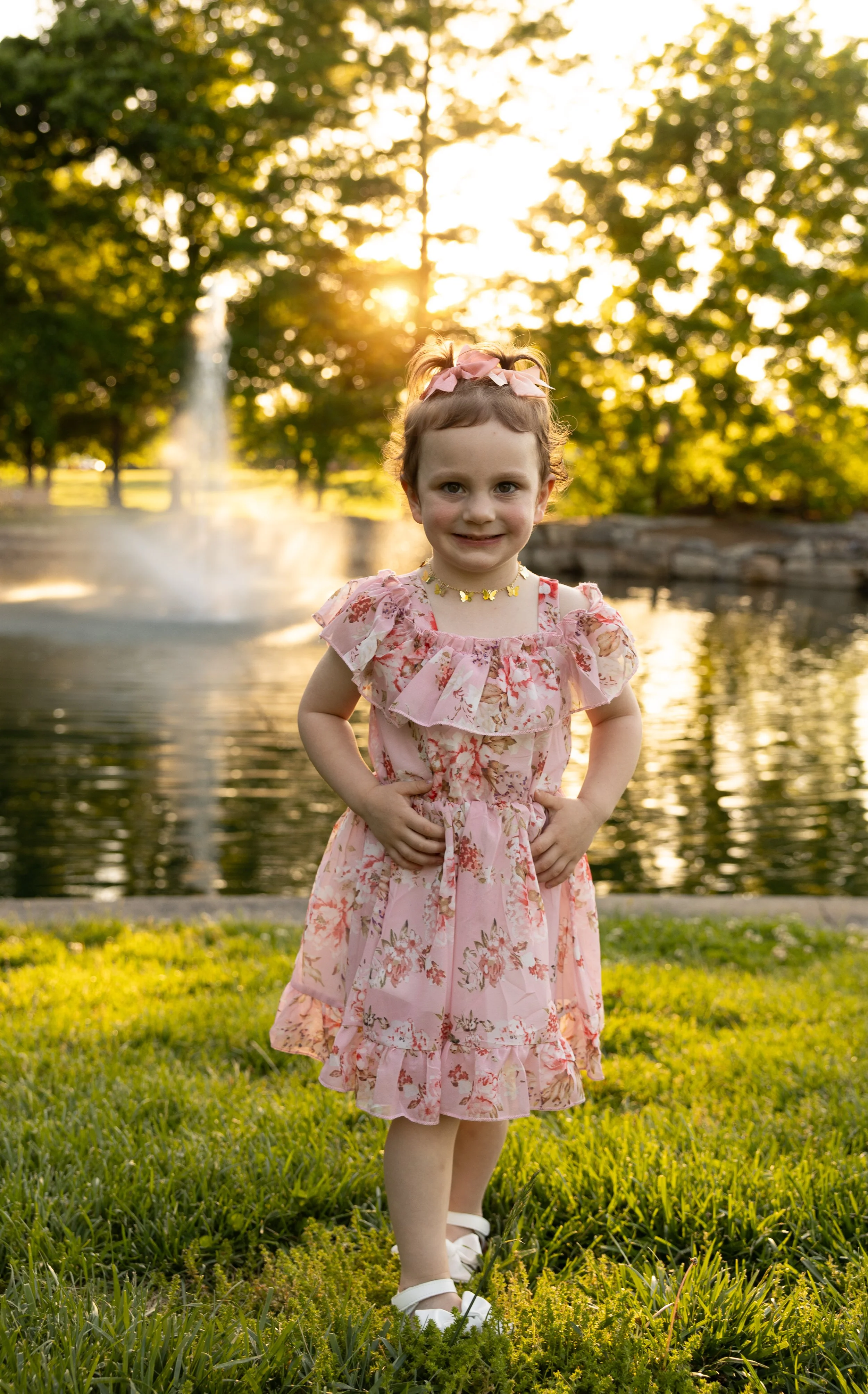 A young girl in a pink floral dress standing on grass by a pond with a fountain and trees in the background, illuminated by the setting sun.