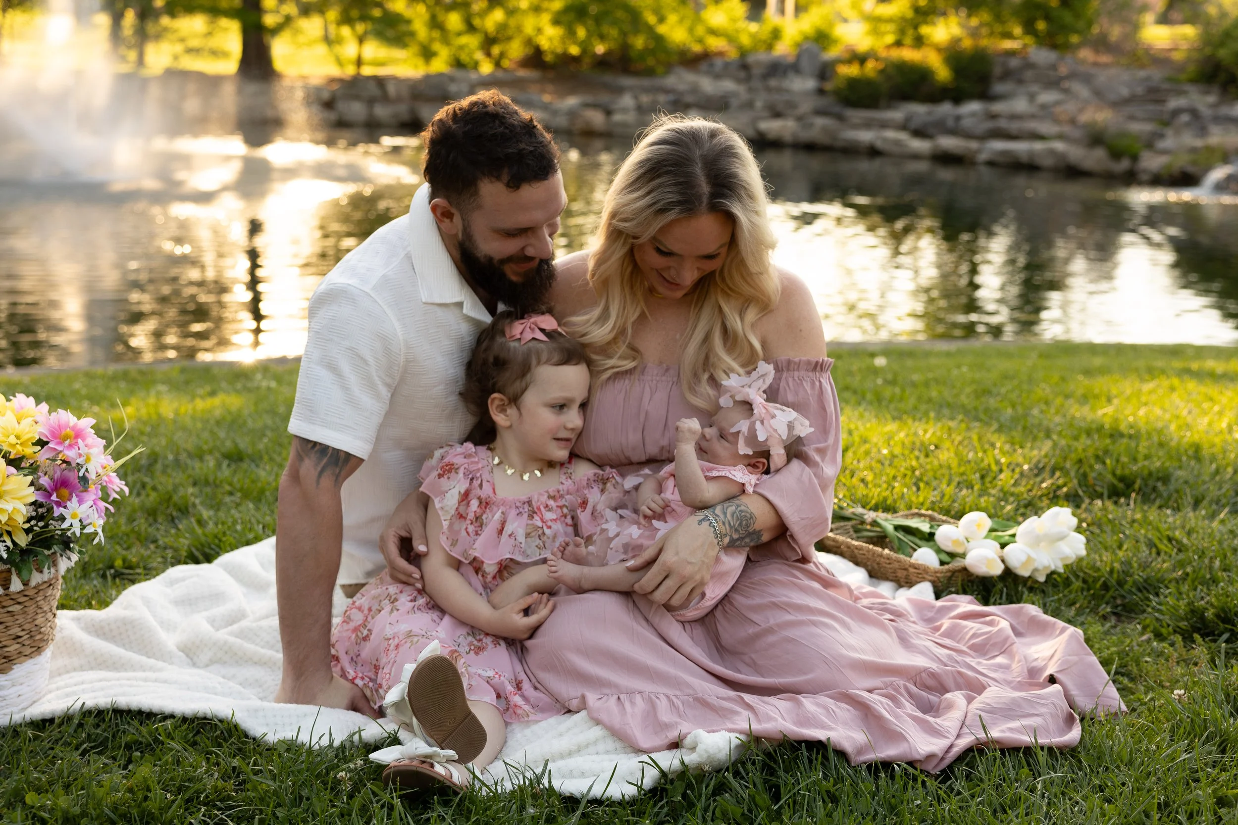 A family of four, including two young girls and parents, sitting on a blanket by a river, smiling and celebrating with flowers and a cake, during sunset.