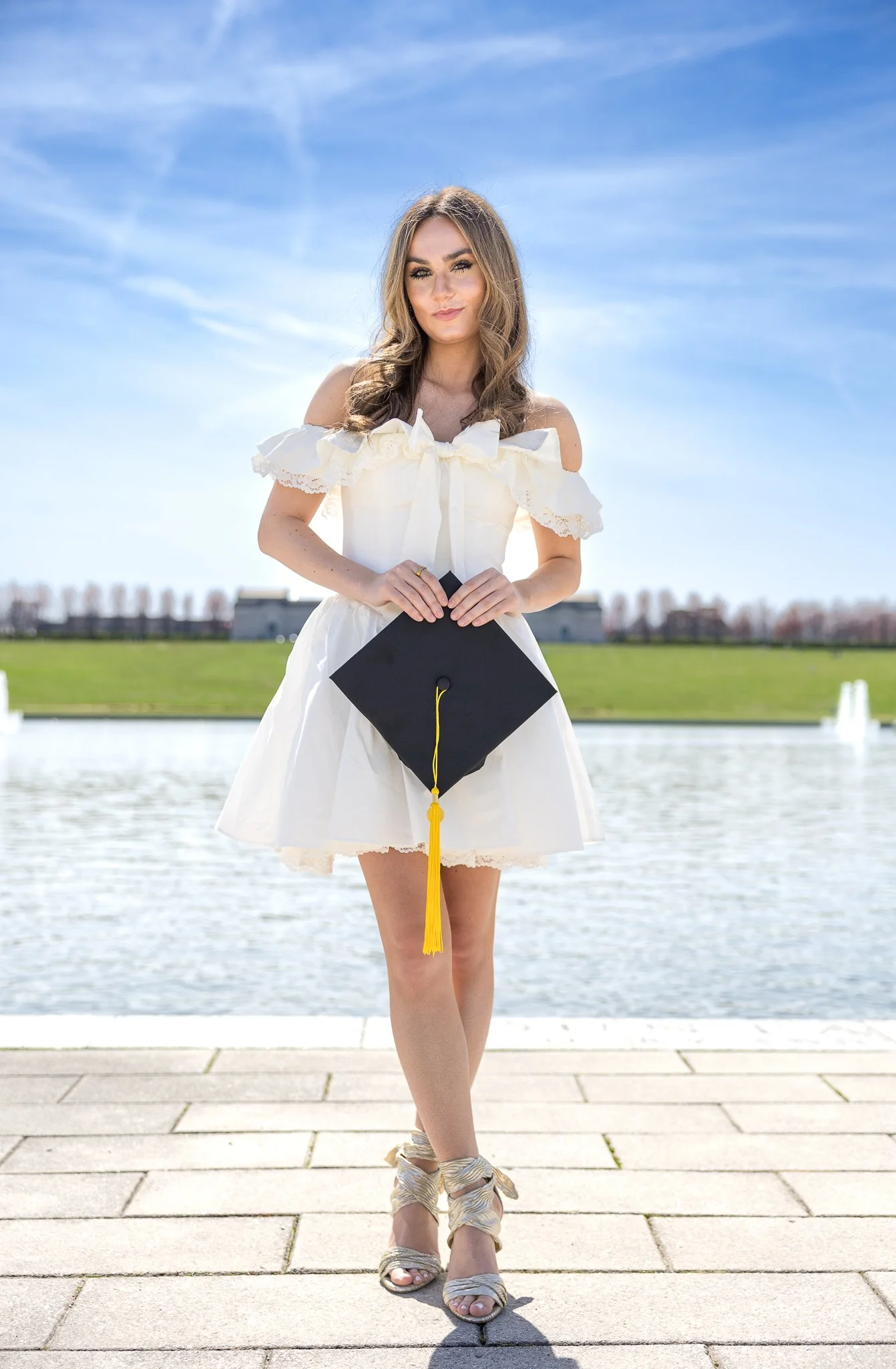 A woman in a white off-the-shoulder dress holding a black graduation cap with a yellow tassel, standing on a paved area near water with a grassy background and a blue sky.