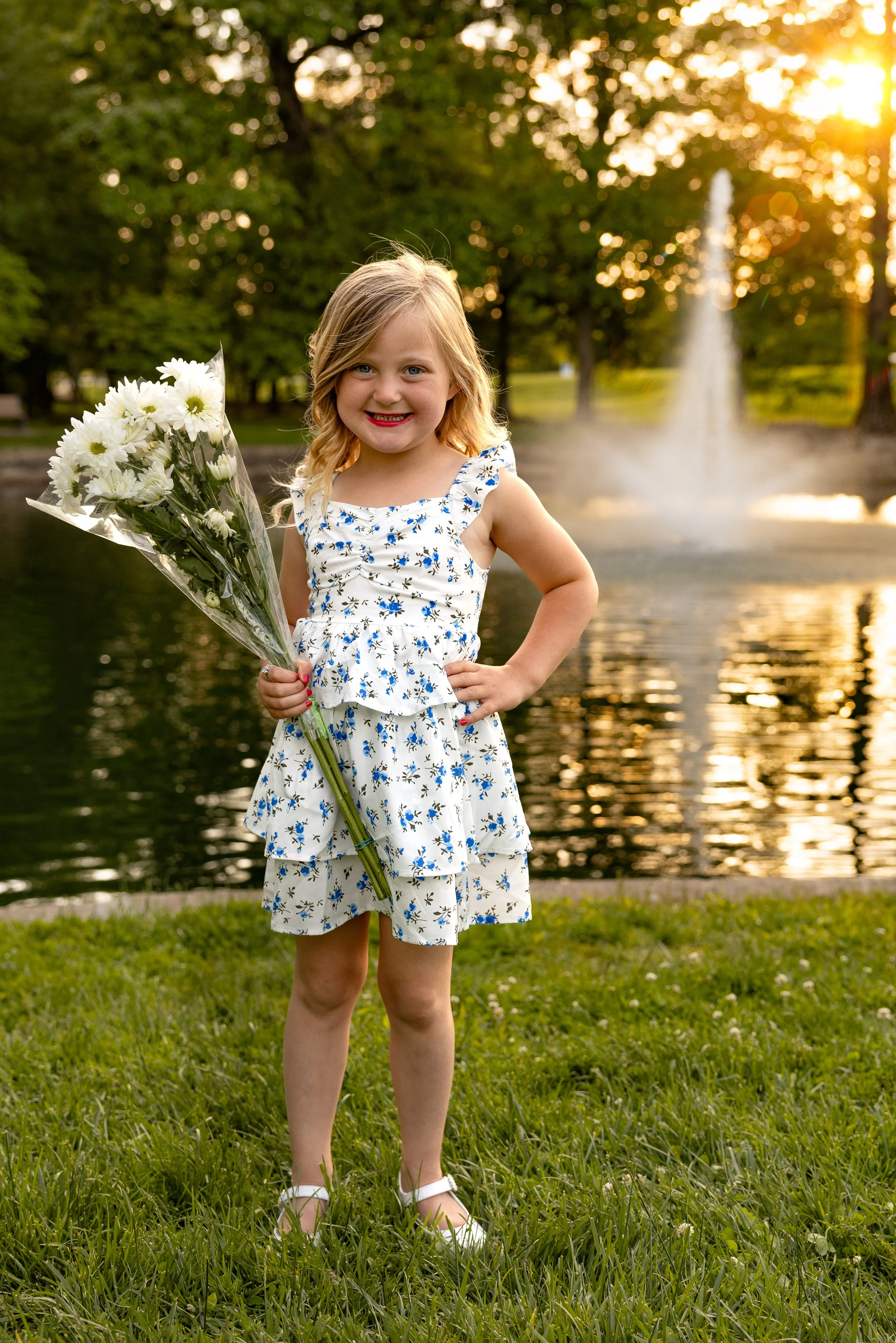 A smiling young girl holding a bouquet of white flowers, standing outdoors by a pond or lake with a fountain, during sunset, wearing a white dress with blue floral pattern, white shoes, and standing on grass.