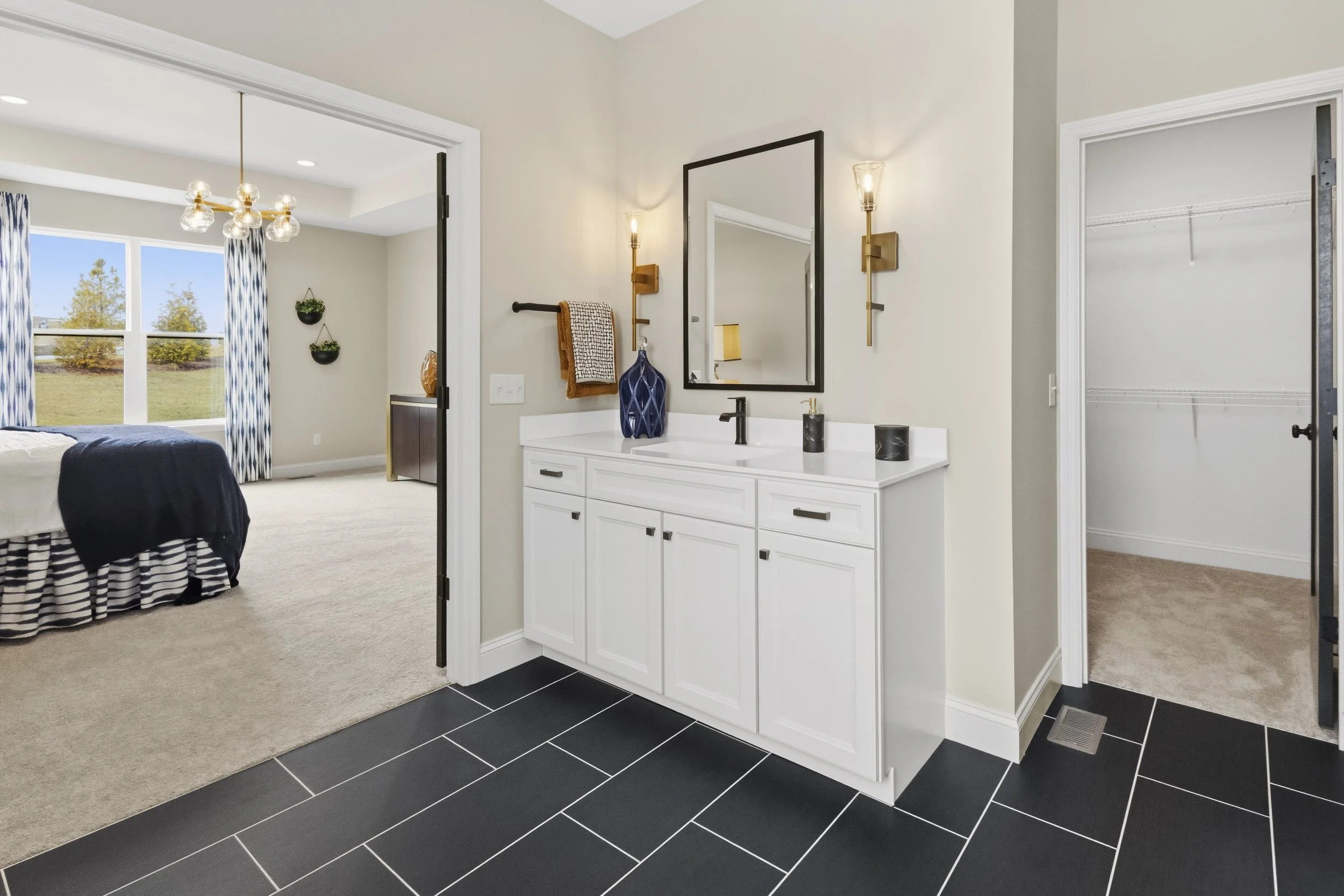 Bathroom vanity with a mirror, two wall sconces, dark faucet, blue vase, and two black containers, with an adjacent bedroom and walk-in closet.