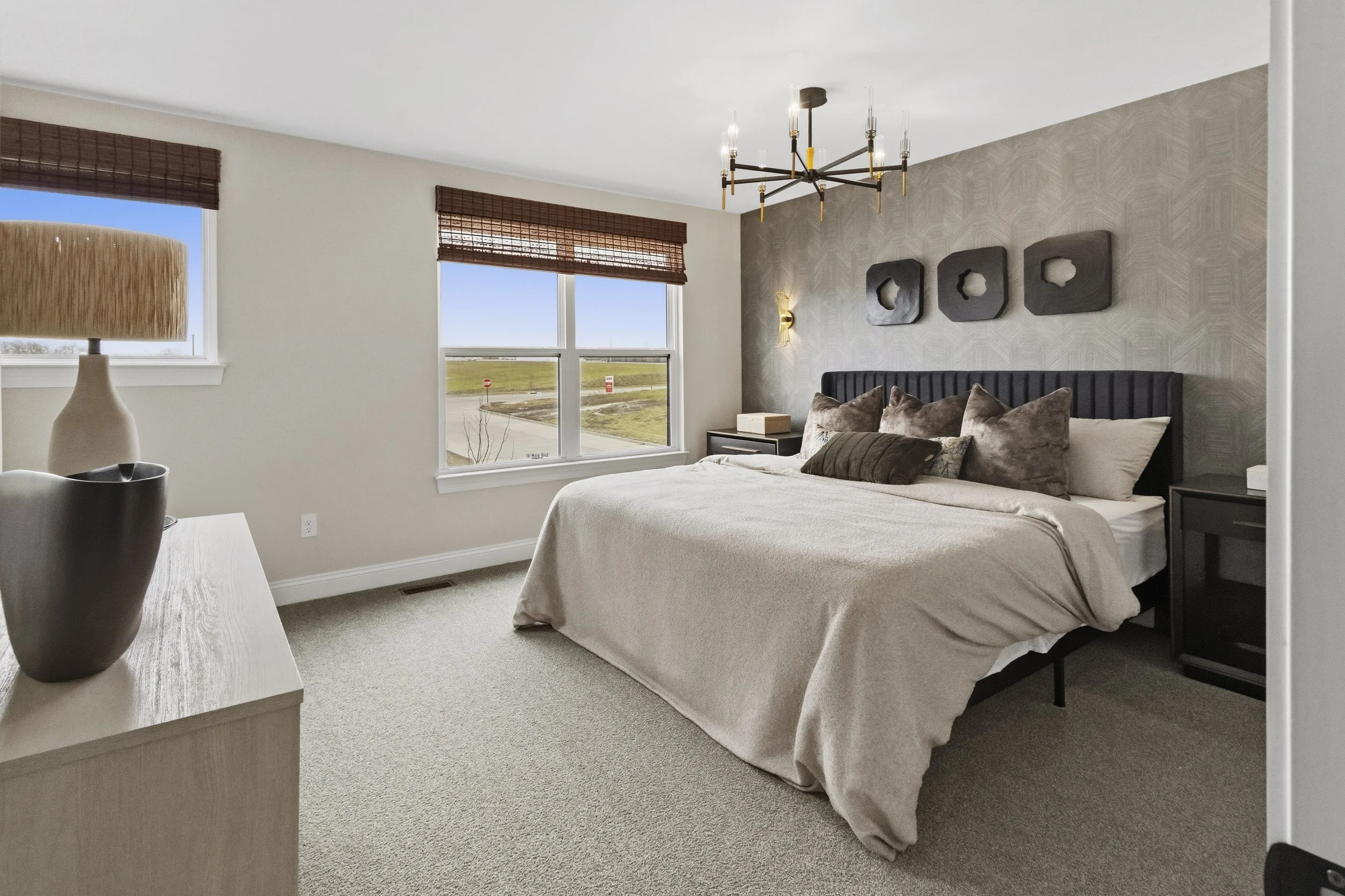 A modern bedroom with a king-sized bed, neutral bedding, and decorative pillows, facing windows with wooden blinds and a view of an open field and a road outside.