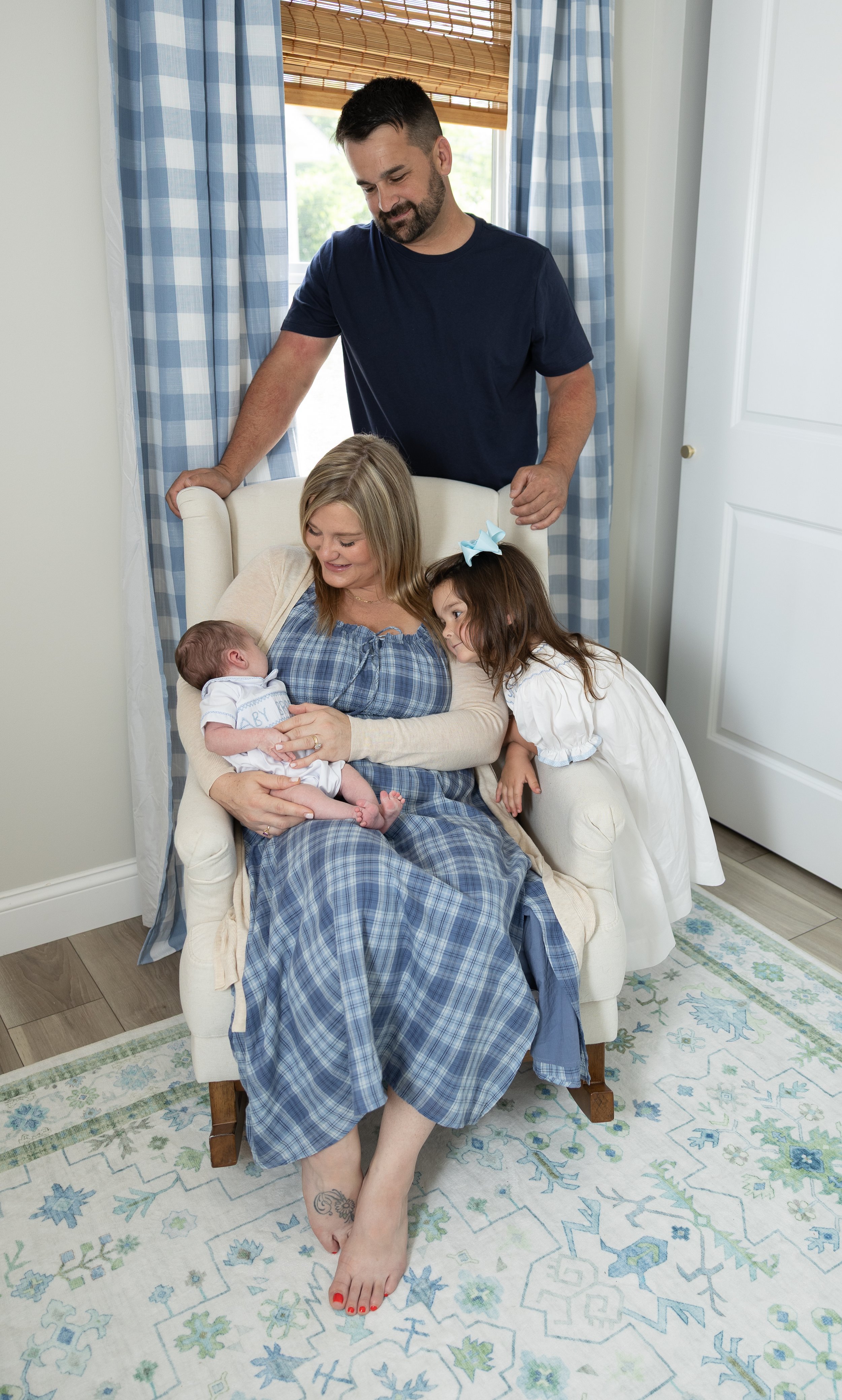 A woman sitting in a white armchair holding a newborn baby with a man standing behind her and a young girl leaning over the chair looking at the baby. The woman is wearing a blue checked dress, and the girl has a white dress with a blue bow in her hair. The man is wearing a dark blue t-shirt. The scene is in a bright room with blue and white checkered curtains and a patterned rug.