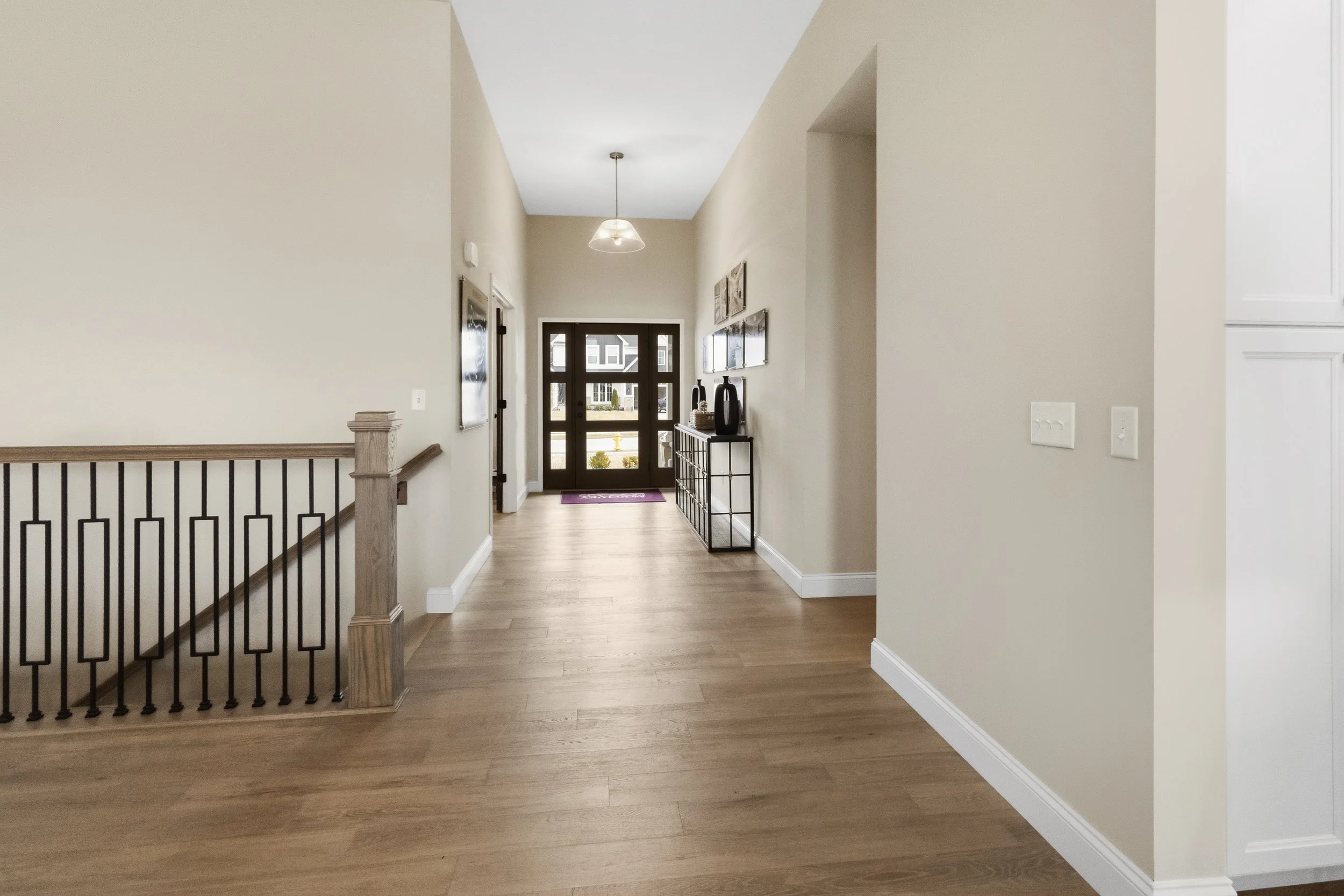 Empty hallway in a modern home with beige walls, wooden flooring, a black front door with glass panels, a ceiling light fixture, and a console table with decorative vases on the right side.