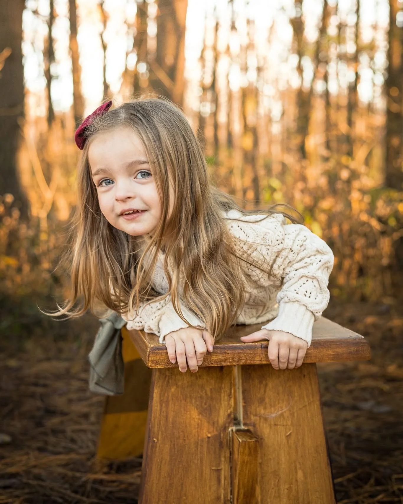 A young girl with long blonde hair, blue eyes, and a pink hair clip, leaning on a wooden bench or table outdoors in a forest during autumn.