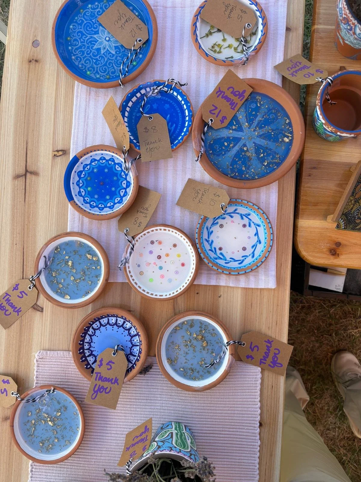 Ceramic dishes and cups on a wooden table, decorated with blue, white, and gold patterns, some with gold leaf accents, with handwritten price and thank you tags attached.