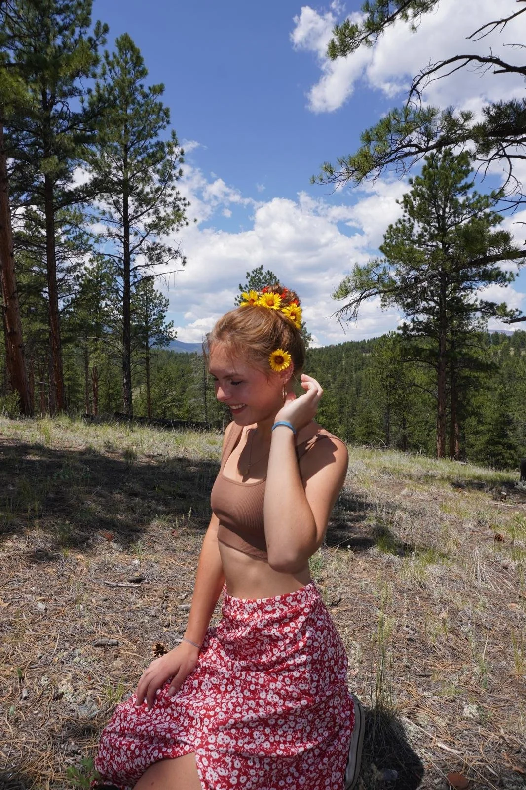 A young woman kneeling outdoors in a forest, decorated with a floral crown and sunflower hair accessories, wearing a beige crop top and a red floral skirt, with a partly cloudy sky overhead.