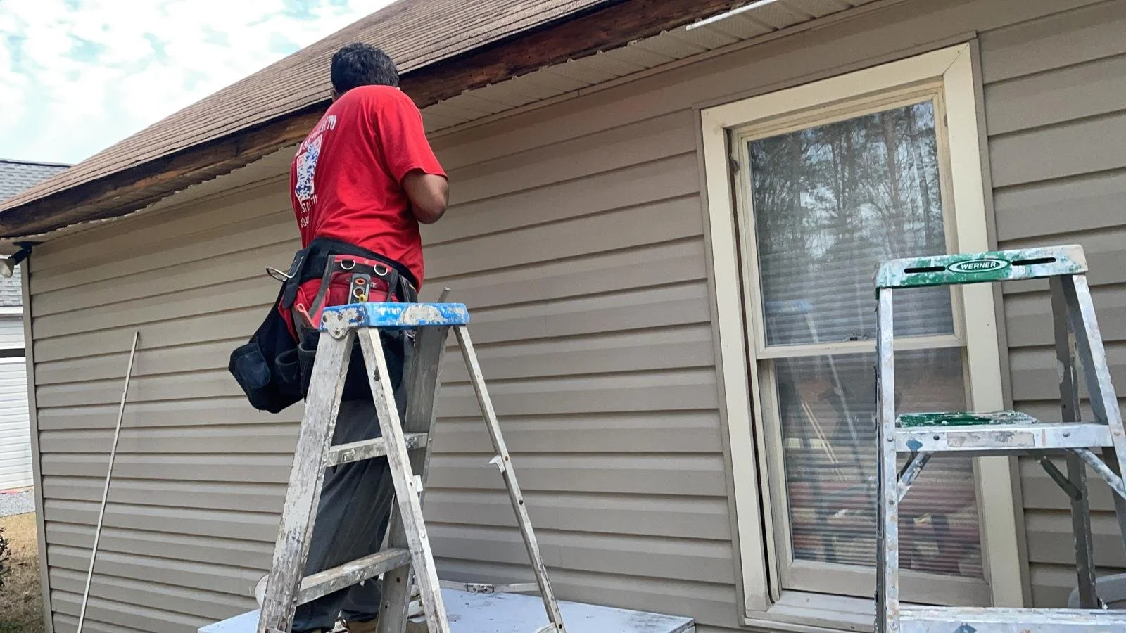 A man in a red shirt standing on a ladder outside a house, working on the roof over a window, with another empty ladder nearby.