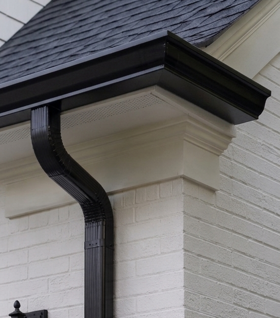 Close-up of a house corner showing a black rain gutter and downspout against a white brick wall and gray shingle roof.