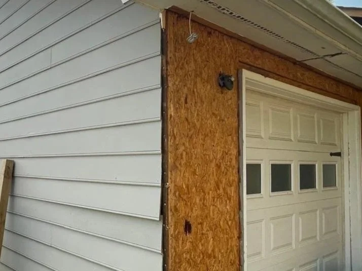 Close-up of a house wall with white vinyl siding and a section of plywood under construction near a garage door.