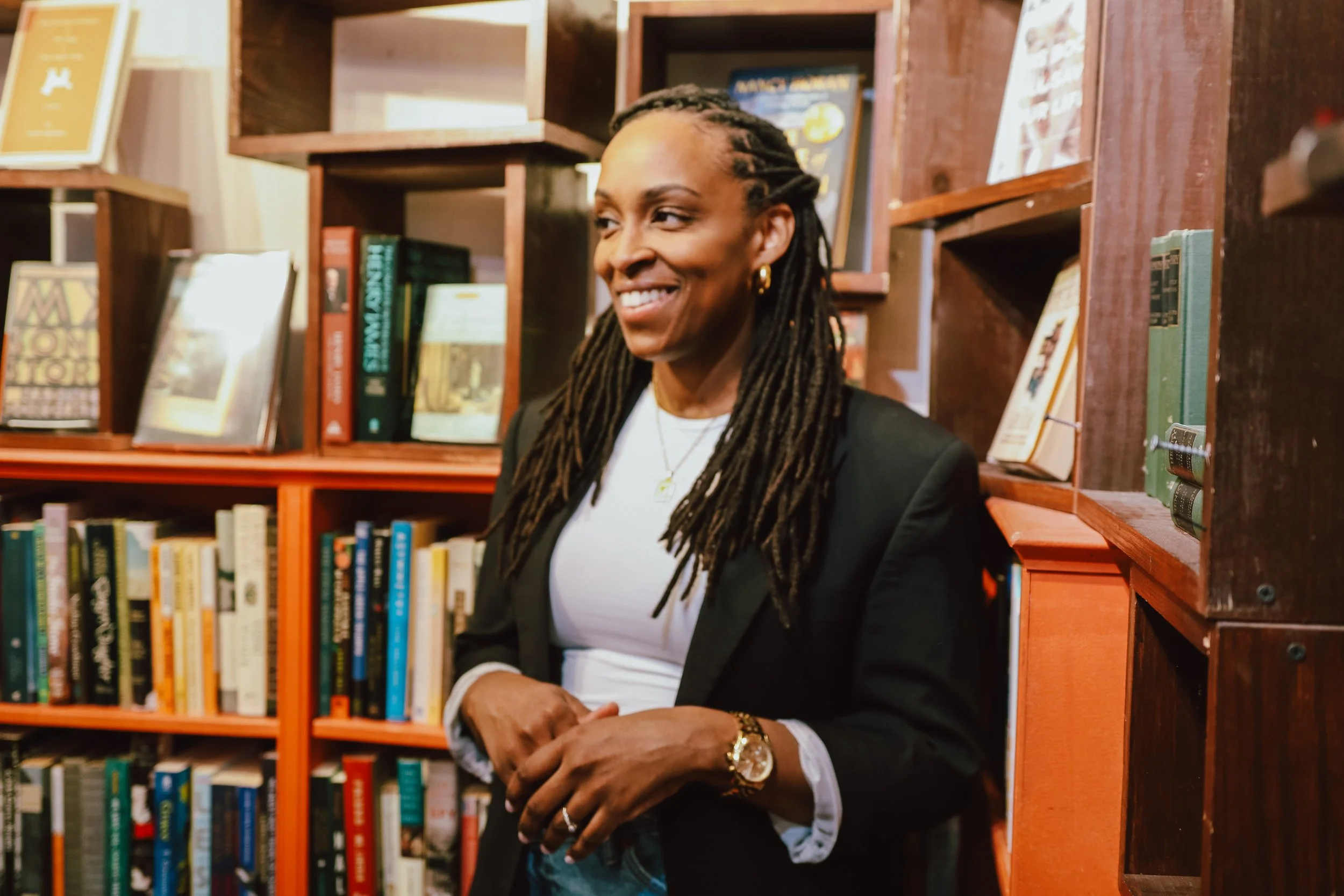 Smiling woman with dreadlocks wearing a black blazer and white shirt standing in front of wooden bookshelves filled with books.