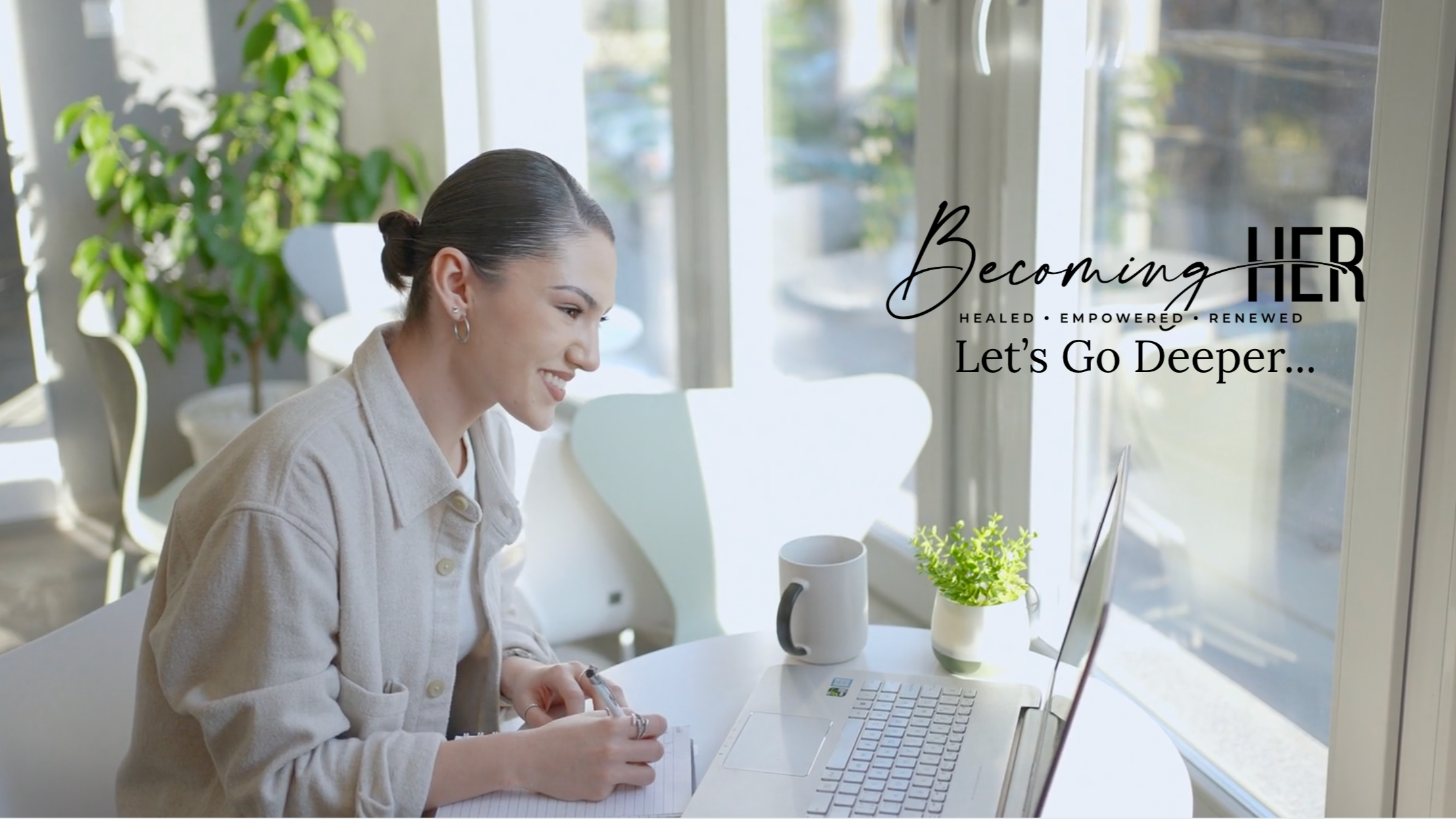 A woman with dark hair tied back, smiling, sitting at a white table near a large window, working on a laptop, with a notebook, pen, and a cup on the table. There are green plants near the window and sunlight streaming in.