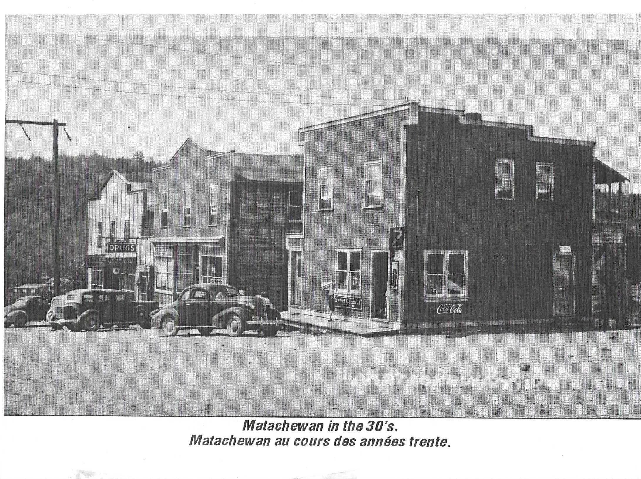 Black and white photo from the 1930s showing a small town street with vintage cars parked in front of buildings. One building has a Coca-Cola sign, another has a sign for a drugstore. The scene includes utility poles and a hill with trees in the background.
