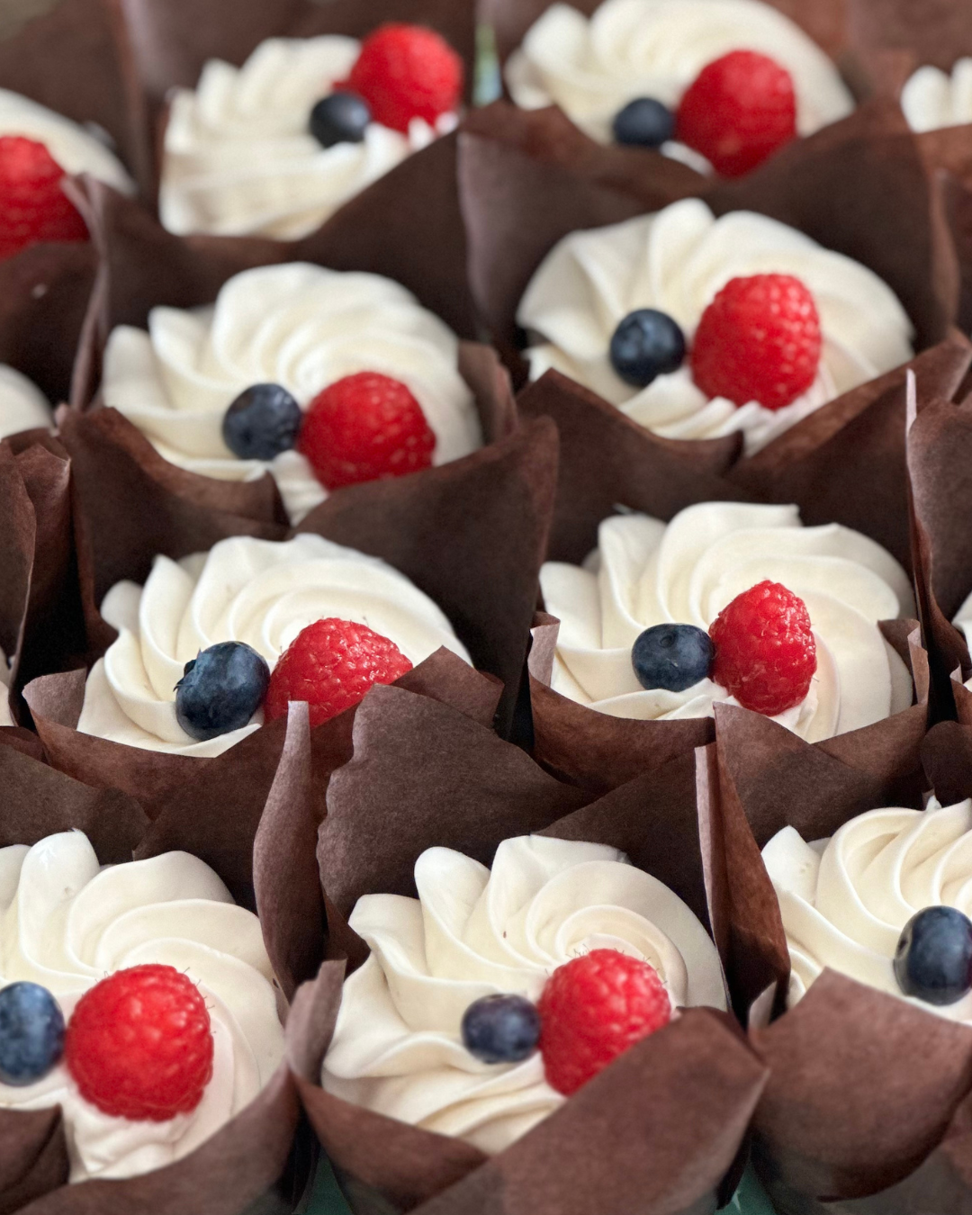 Cupcakes with white frosting topped with strawberries and blueberries, wrapped in brown paper wrappers.
