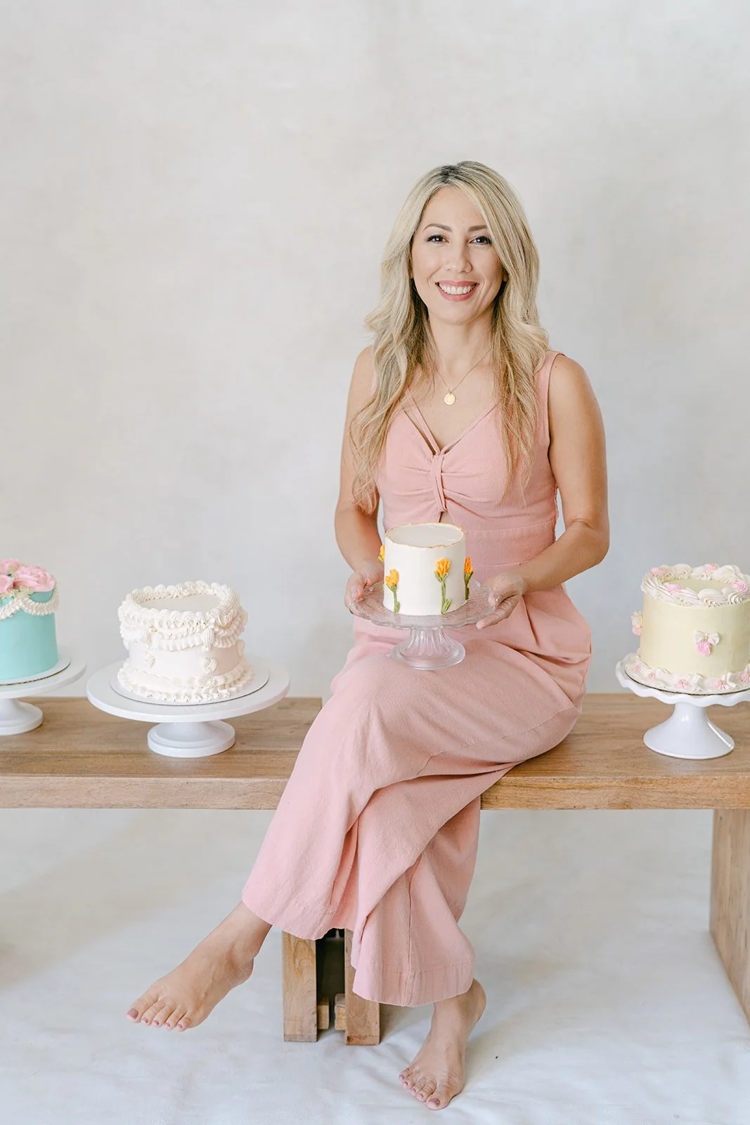 Woman in pink dress sitting on a wooden bench holding a decorated cake, with three more decorated cakes on stands nearby.