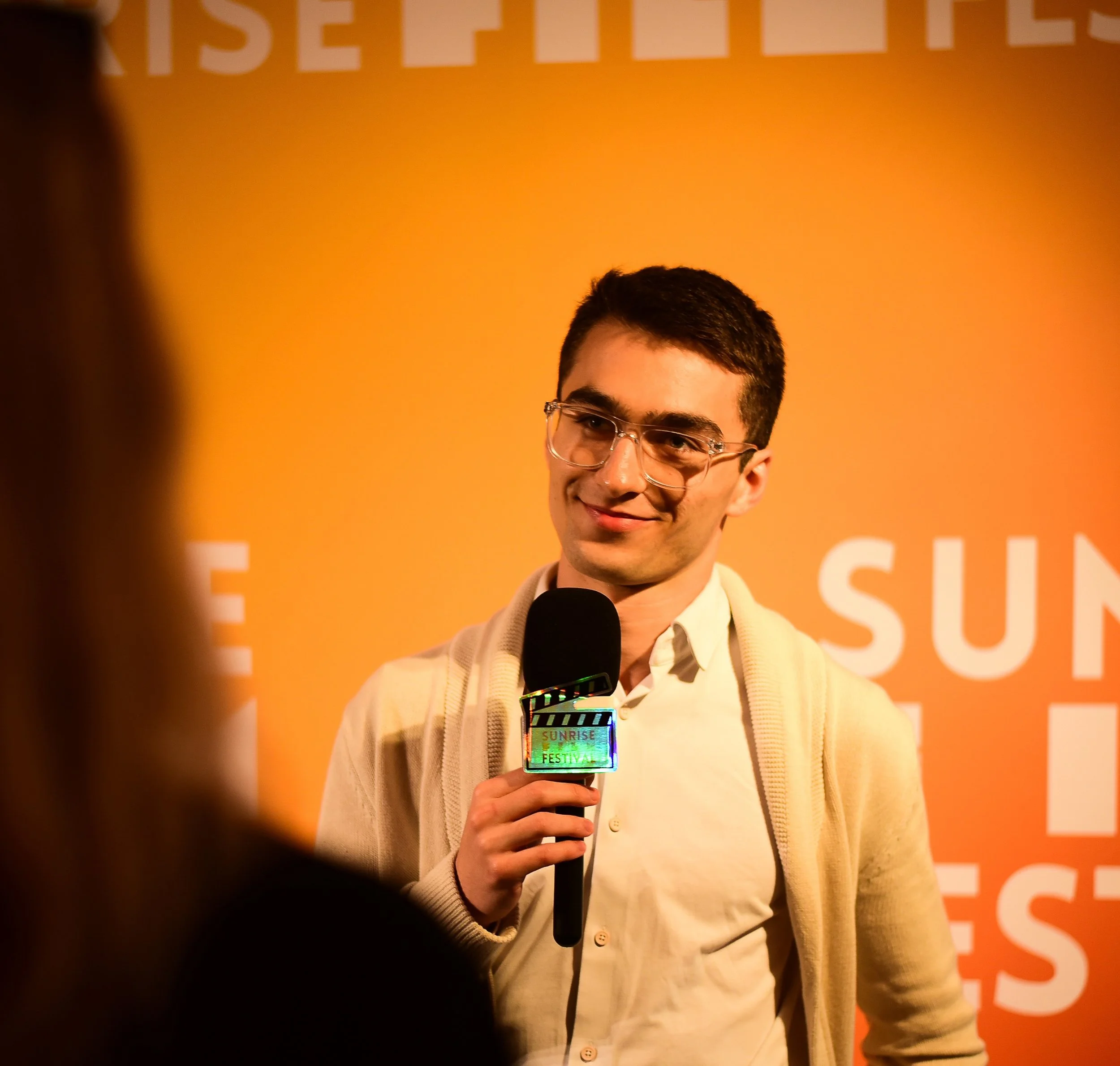 A young man with dark hair and glasses holding a microphone at a Sunrise Festival event, with an orange background and partial white text.