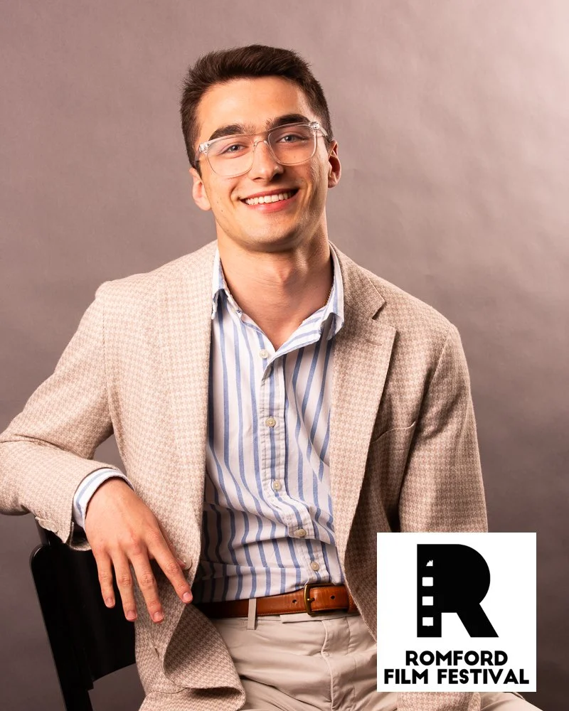 A young man with short dark hair and glasses, smiling, dressed in a beige blazer and striped shirt, sitting on a black chair with a gray background. Romford Film Festival logo in the bottom right corner.
