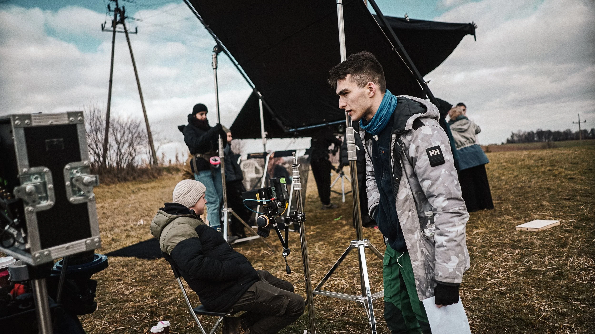 Film crew set up equipment outdoors on a cloudy day, with a man sitting on a chair and a crew member standing nearby.