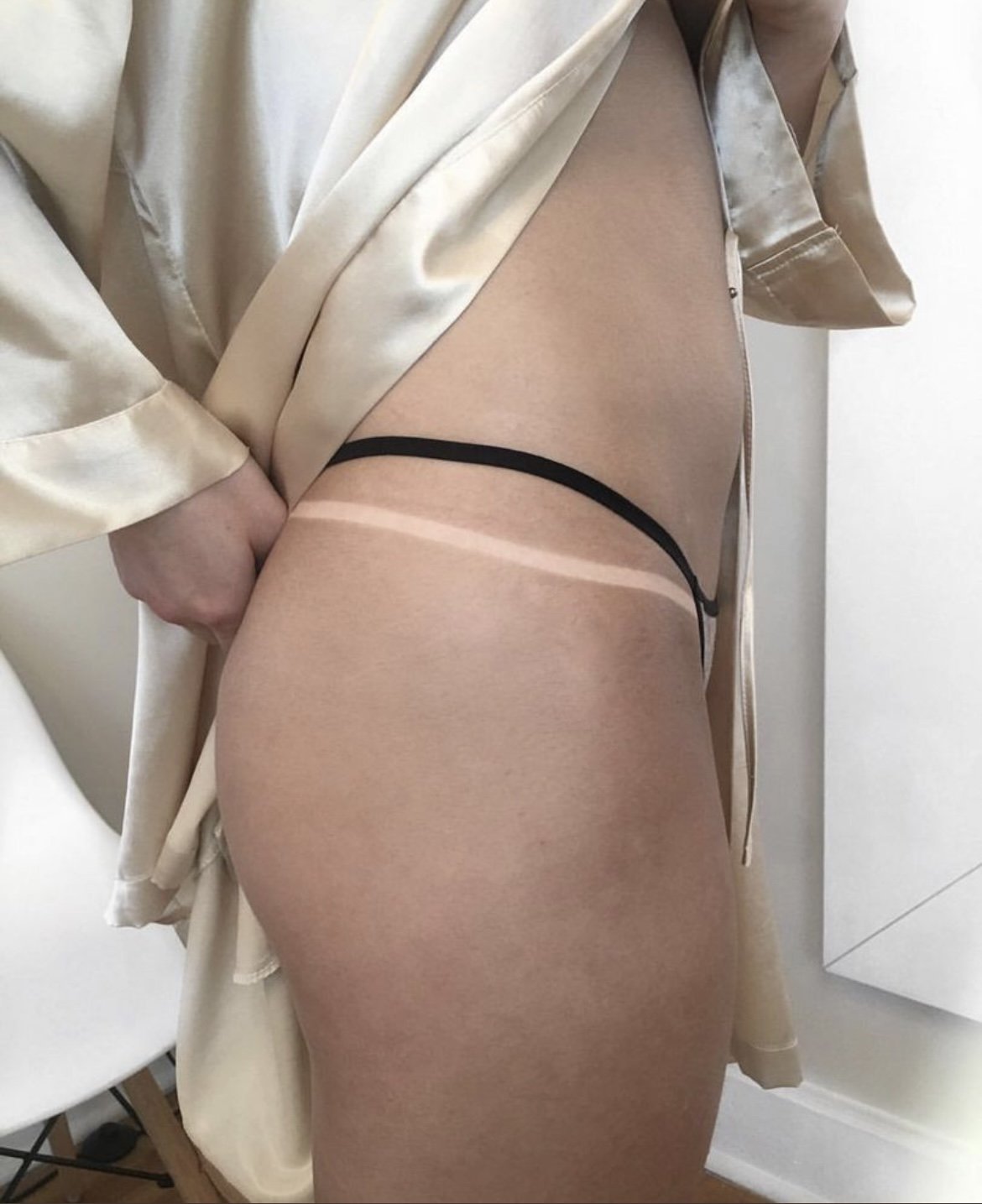 A person wearing a beige satin robe, black thong, and nude stockings is pulling up the robe to reveal their thigh, buttocks, and the top of the stocking line.