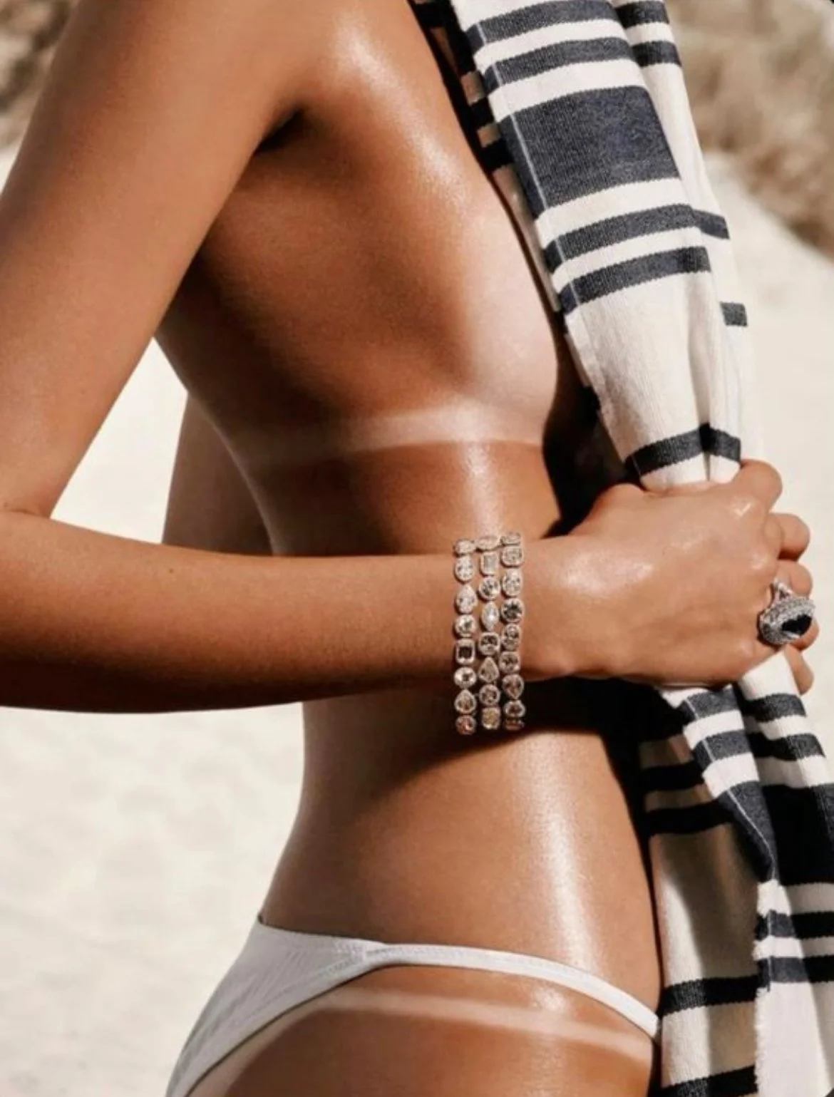 Close-up of a woman with tanned skin, holding a black and white striped towel, wearing white underwear, and multiple silver bracelets on her wrist.