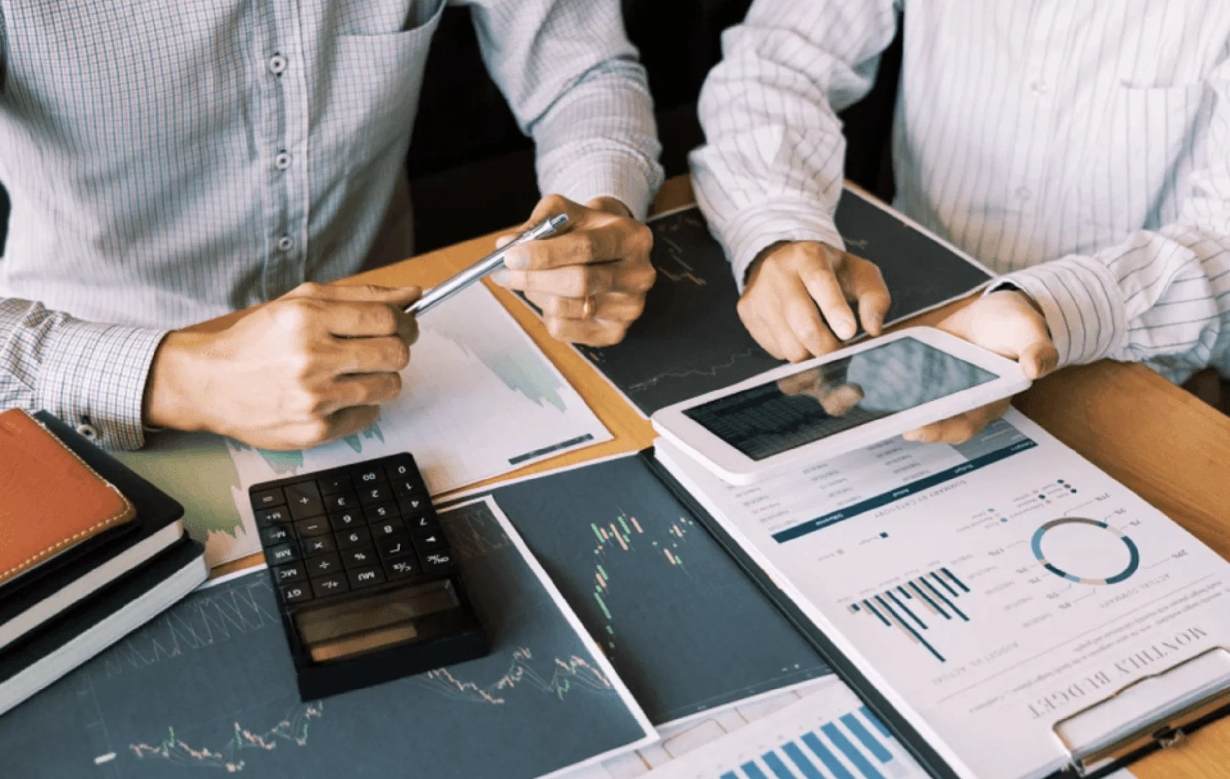 Two people analyzing financial data using a tablet, smartphone, and printed charts at a wooden desk.