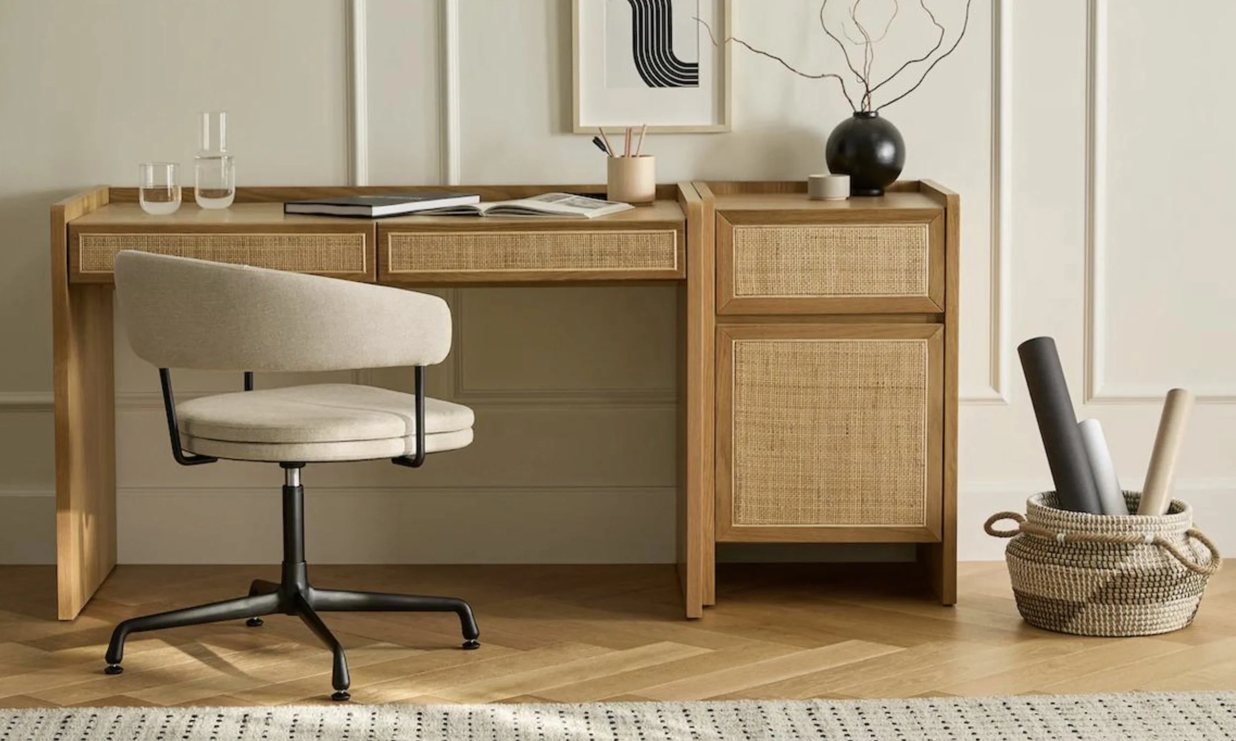A beige office chair in front of a wooden desk with two drawers and woven cabinet doors. On top of the desk are two glasses of water, a closed book, an open book, and a small plant holder with brushes. To the right of the desk is a woven basket with rolled-up papers or posters. The background features wall paneling and framed abstract art.