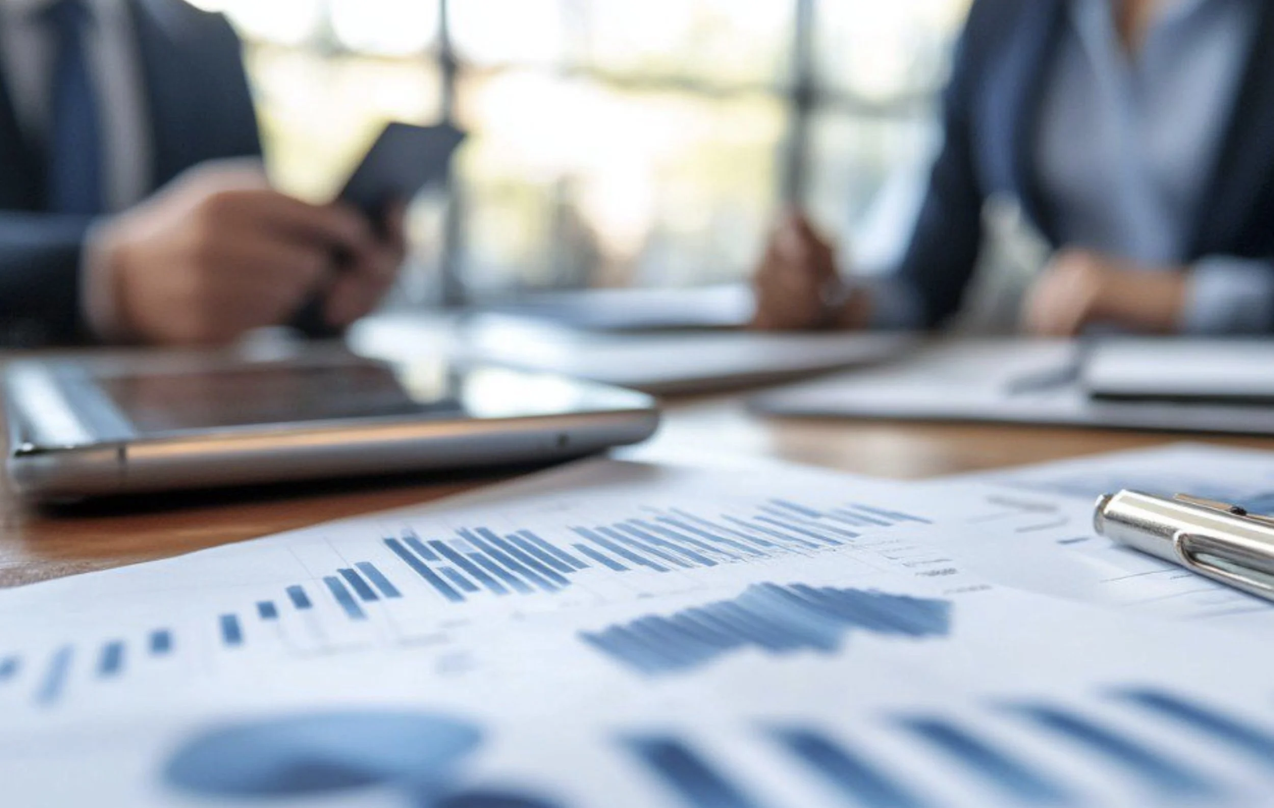 Business meeting with two people in suits sitting at a conference table with documents, a tablet, a smartphone, and a pen, with blurred background