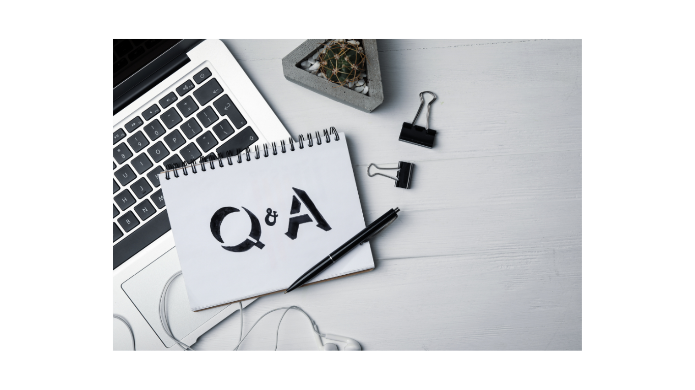 A laptop computer, a notepad with 'Q & A' written on it, a pen, a small potted cactus, and two black binder clips on a white wooden desk.